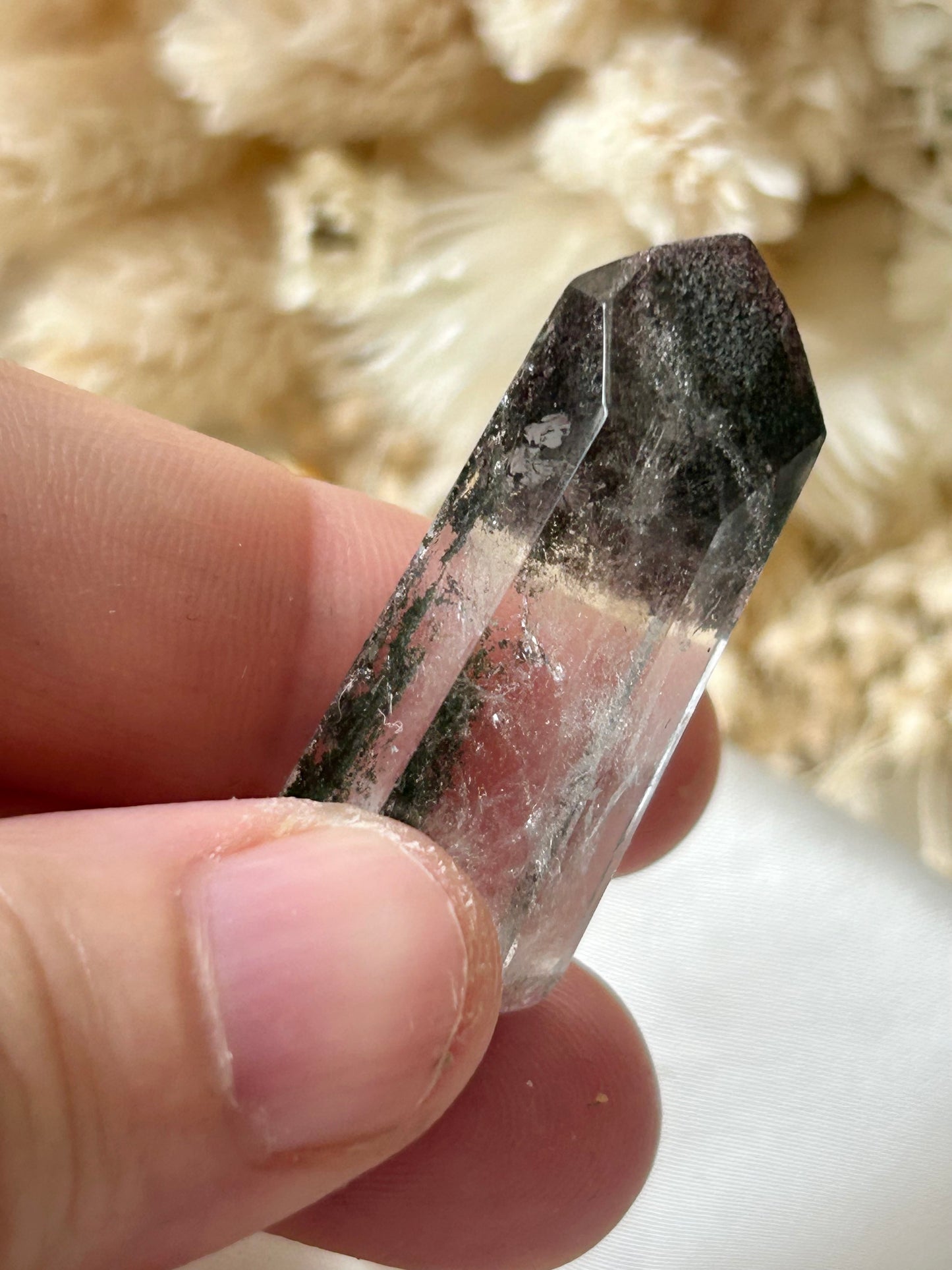 holding a Lodolite freeform crystal against an ivory satin backdrop with dried cream flowers; soft natural light highlights internal inclusions.