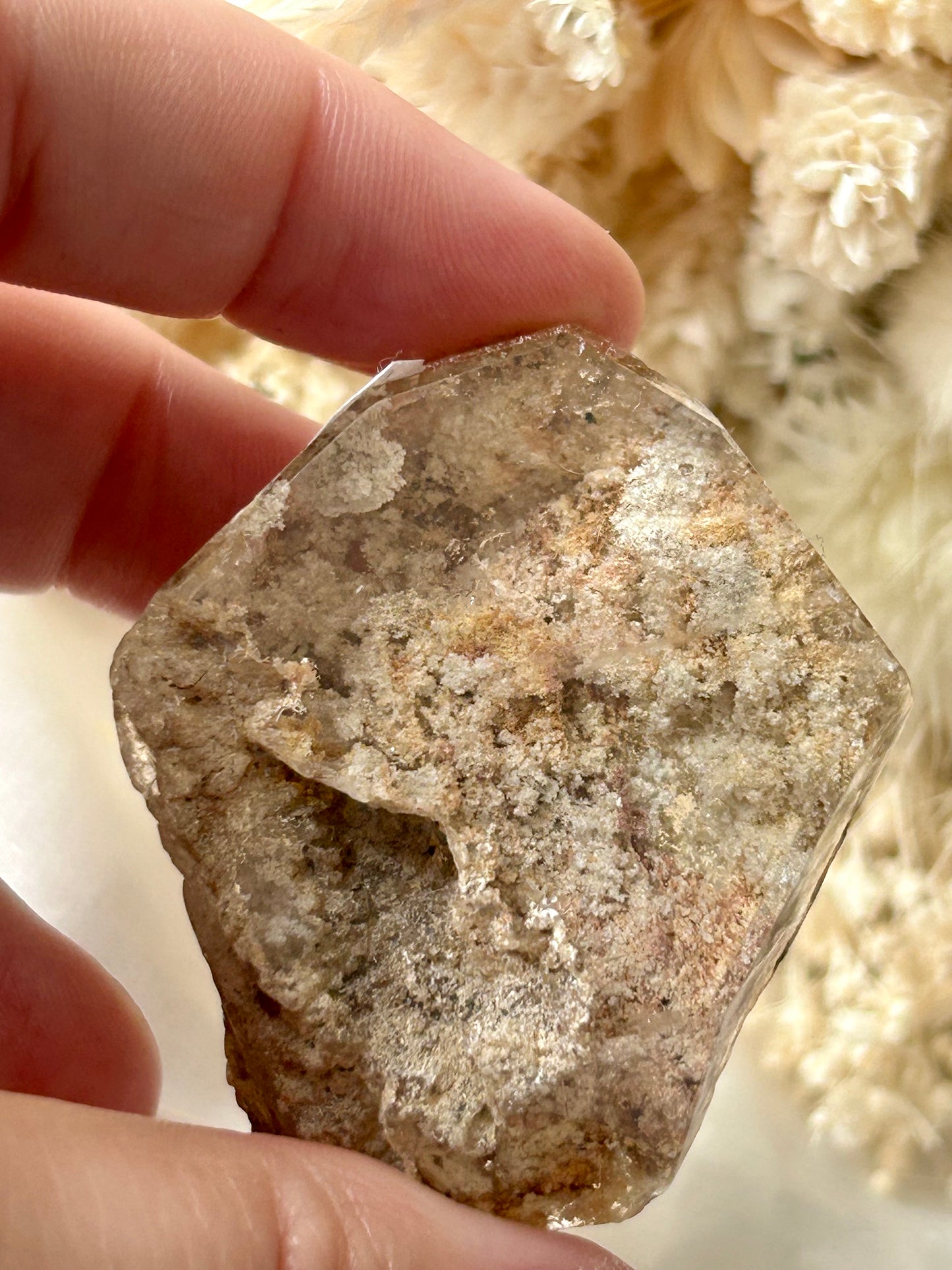holding a Lodolite freeform crystal against an ivory satin backdrop with dried cream flowers; soft natural light highlights internal inclusions.
