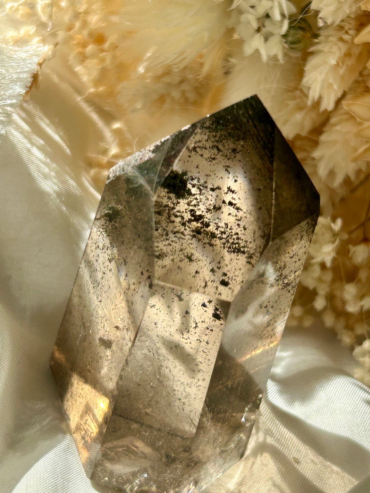 Smoky Quartz tower with chlorite inclusions resting on ivory satin, catching golden sunlight with earthy internal reflections.