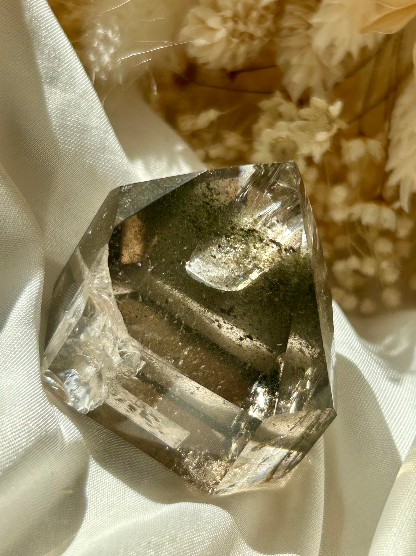 Smoky Quartz tower with chlorite inclusions resting on ivory satin, catching golden sunlight with earthy internal reflections.