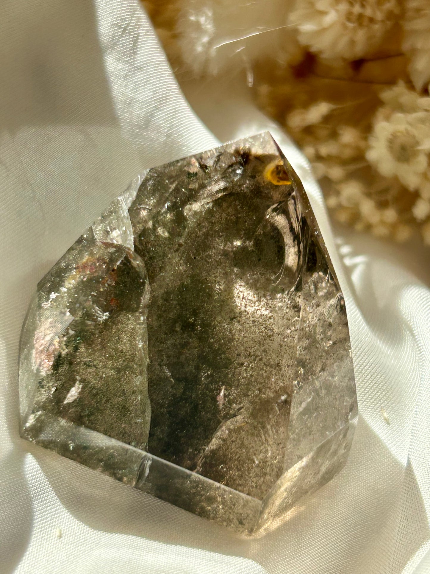 Smoky Quartz tower with chlorite inclusions resting on ivory satin, catching golden sunlight with earthy internal reflections.