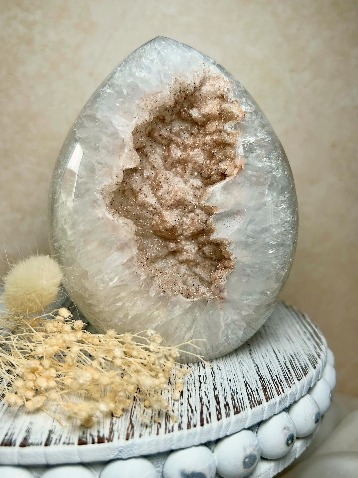 Rainbow Amethyst geode egg with shimmering druzy interior and soft pastel sparkle, displayed on a rustic white stand beside dried florals.