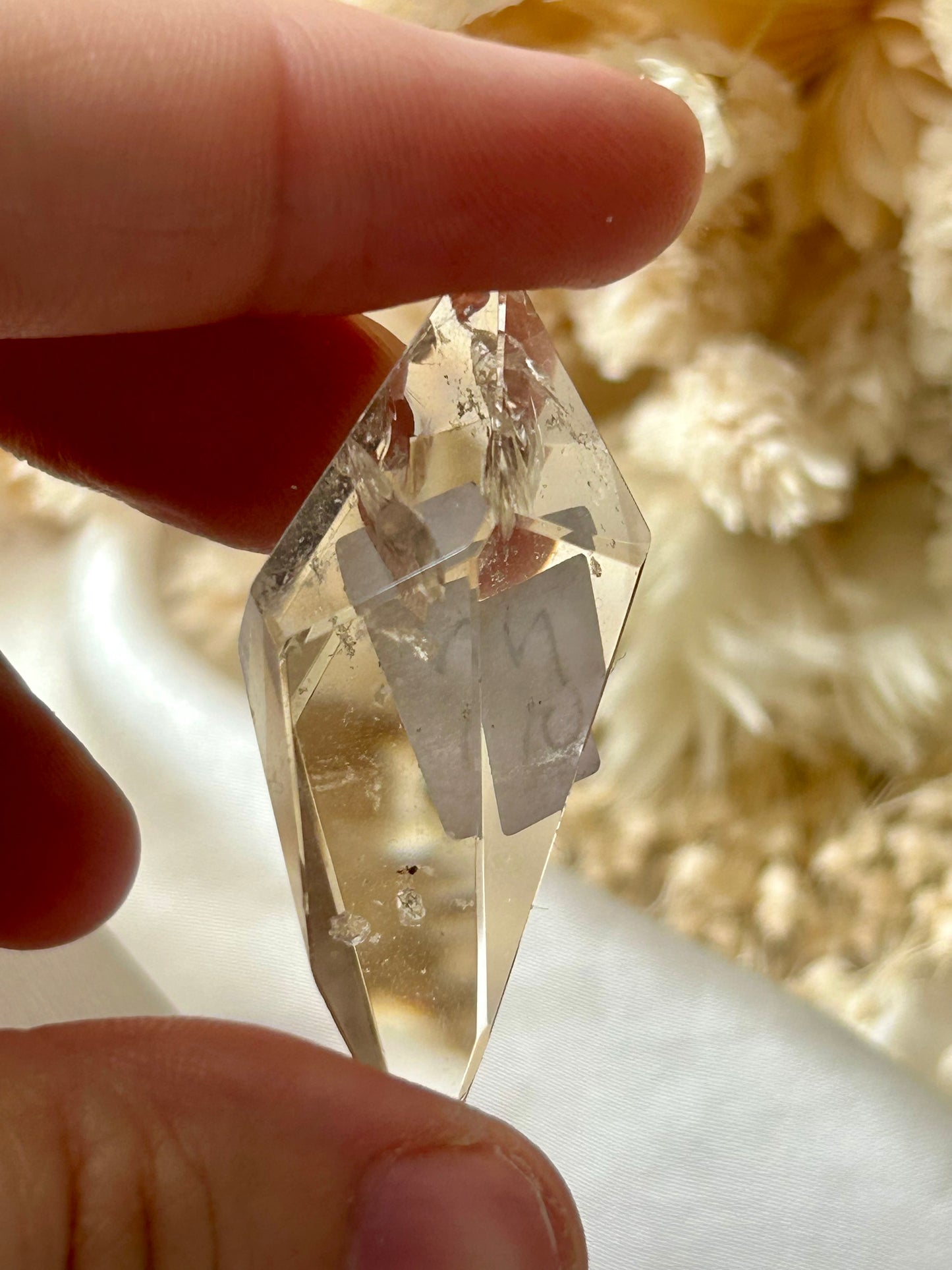 holding a Lodolite freeform crystal against an ivory satin backdrop with dried cream flowers; soft natural light highlights internal inclusions.