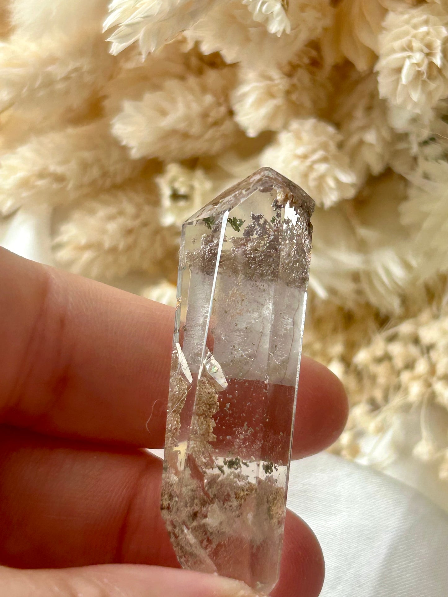 holding a Lodolite freeform crystal against an ivory satin backdrop with dried cream flowers; soft natural light highlights internal inclusions.