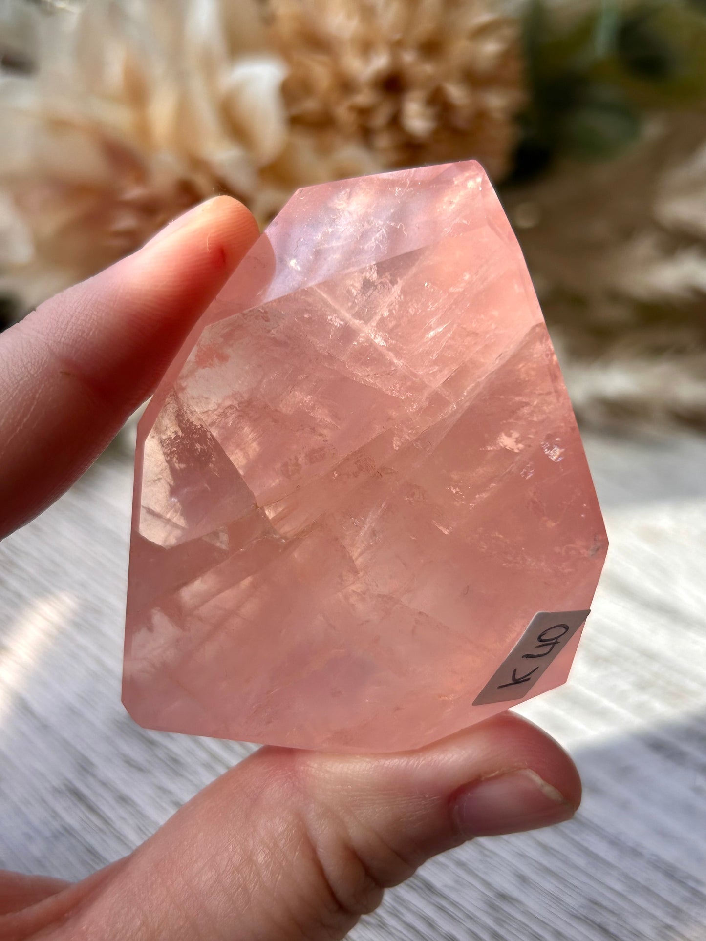 A hand holding a rough, faceted pink crystal stone with a small handwritten label attached near the bottom right. The background is a soft-focus indoor setting with neutral dried flowers and light-colored surfaces.