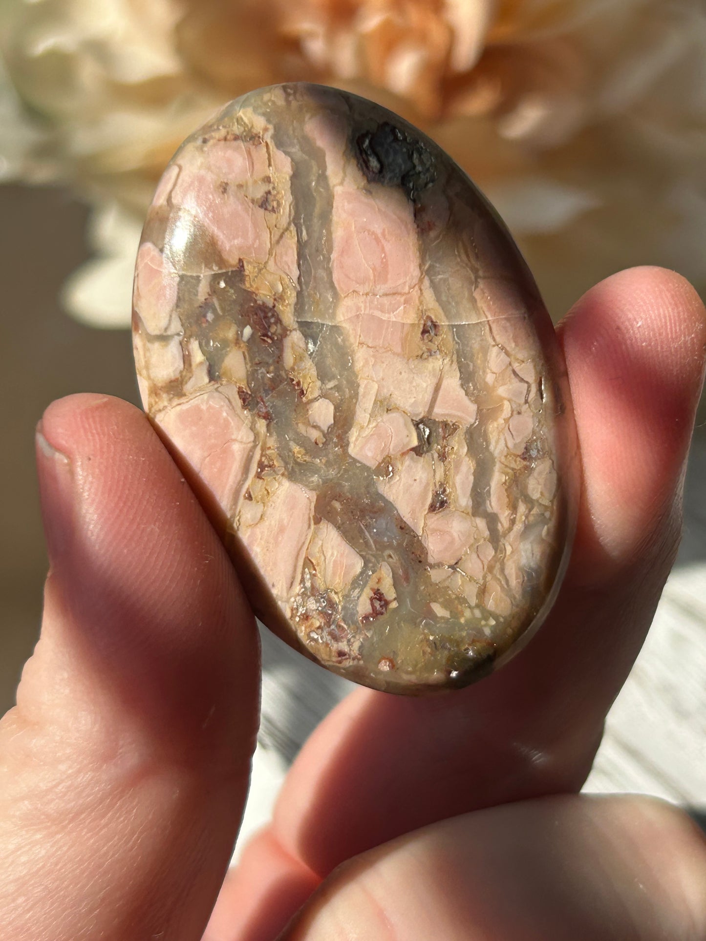 A hand holding a polished oval cabochon stone with a natural marbled pattern of pink, beige, and gray.