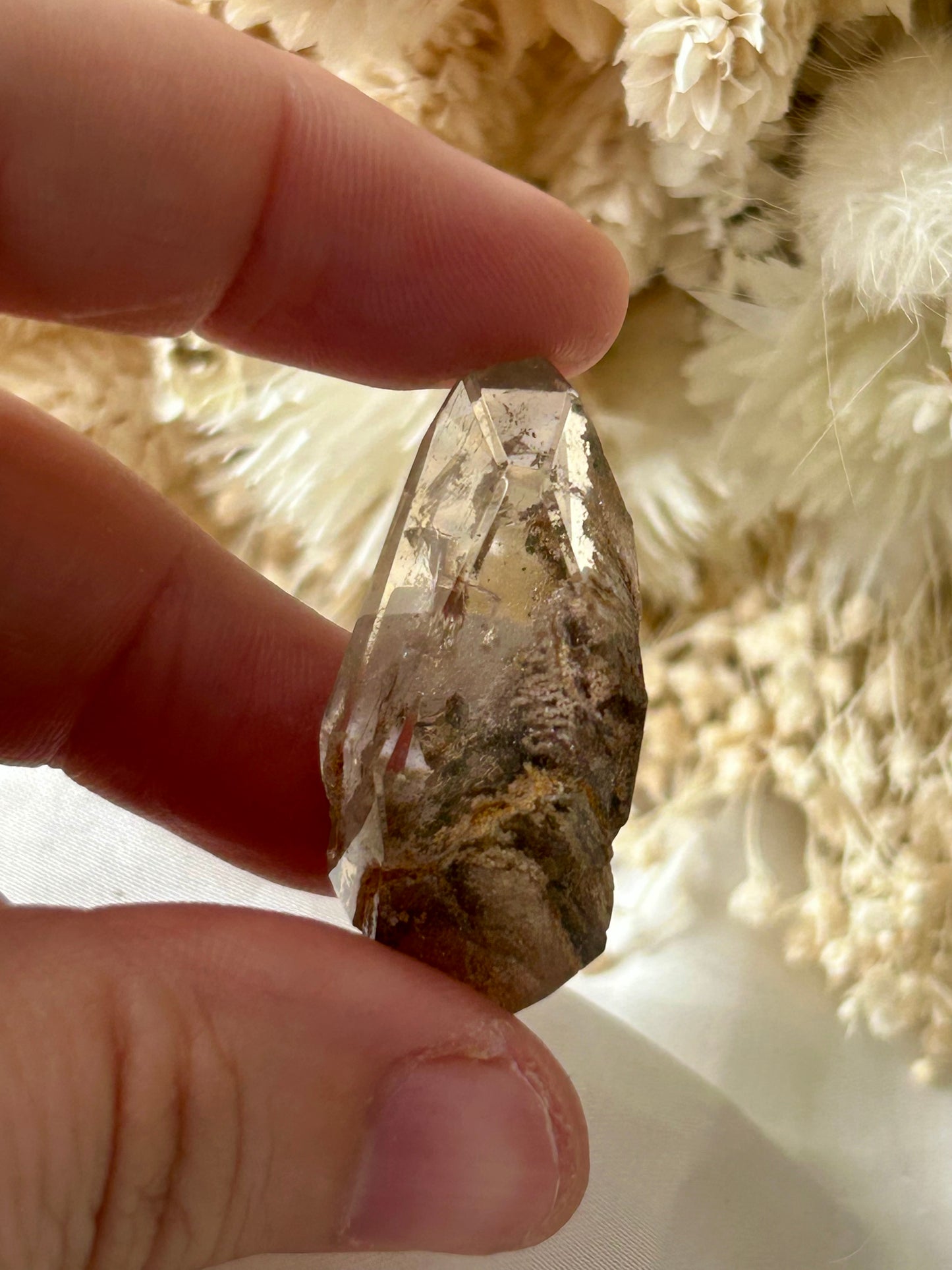 holding a Lodolite freeform crystal against an ivory satin backdrop with dried cream flowers; soft natural light highlights internal inclusions.