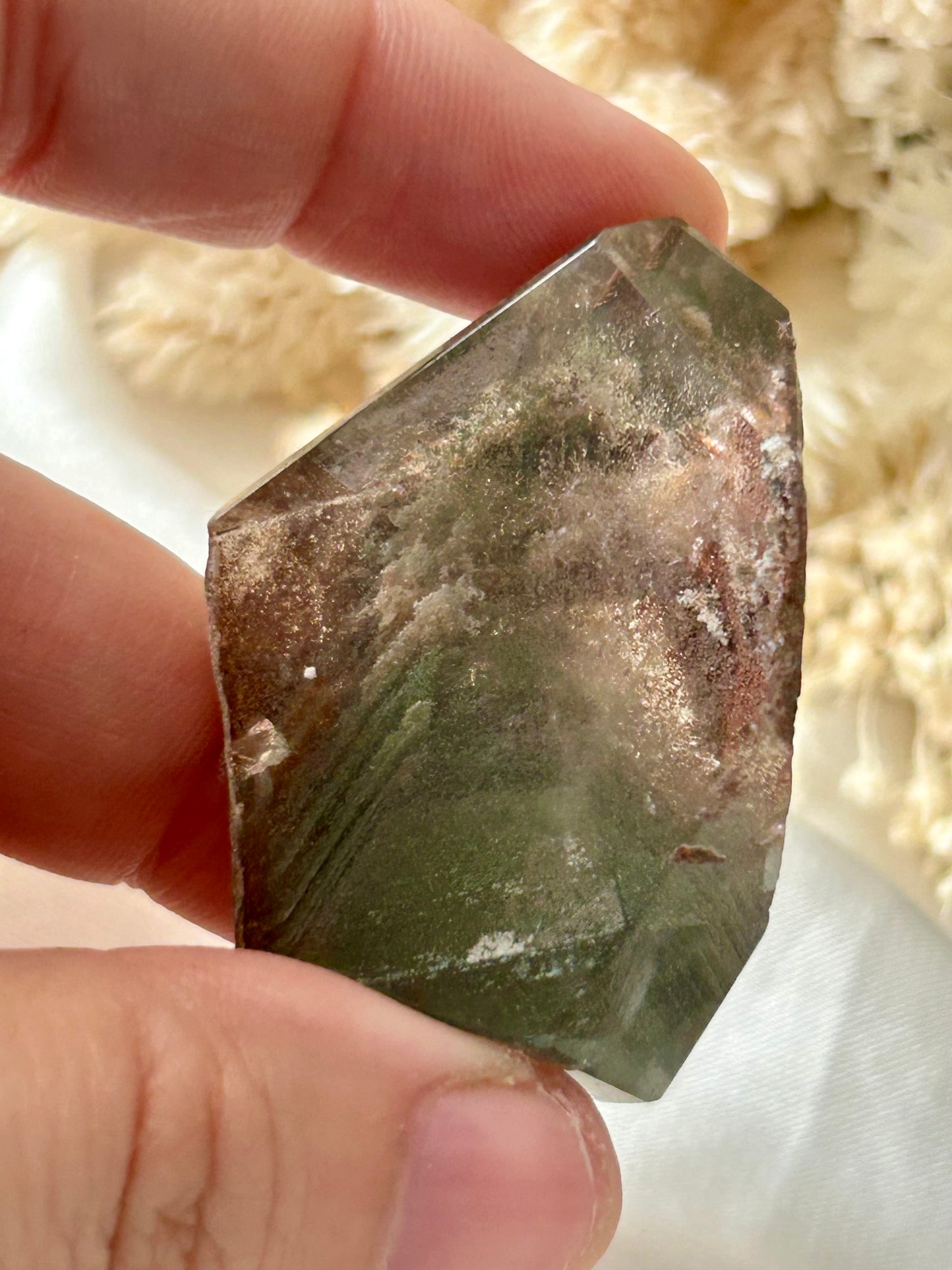 holding a Lodolite freeform crystal against an ivory satin backdrop with dried cream flowers; soft natural light highlights internal inclusions.