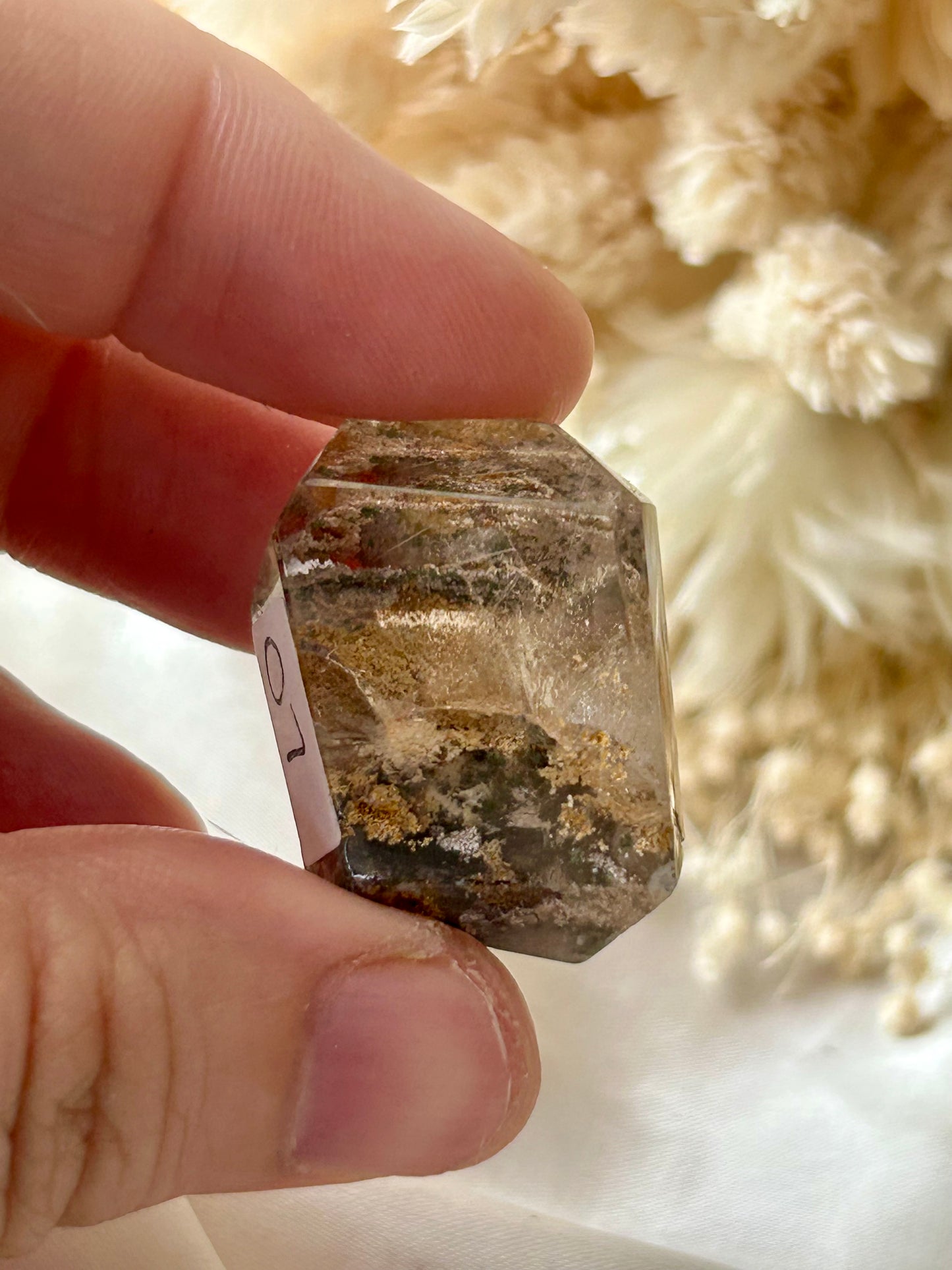 holding a Lodolite freeform crystal against an ivory satin backdrop with dried cream flowers; soft natural light highlights internal inclusions.