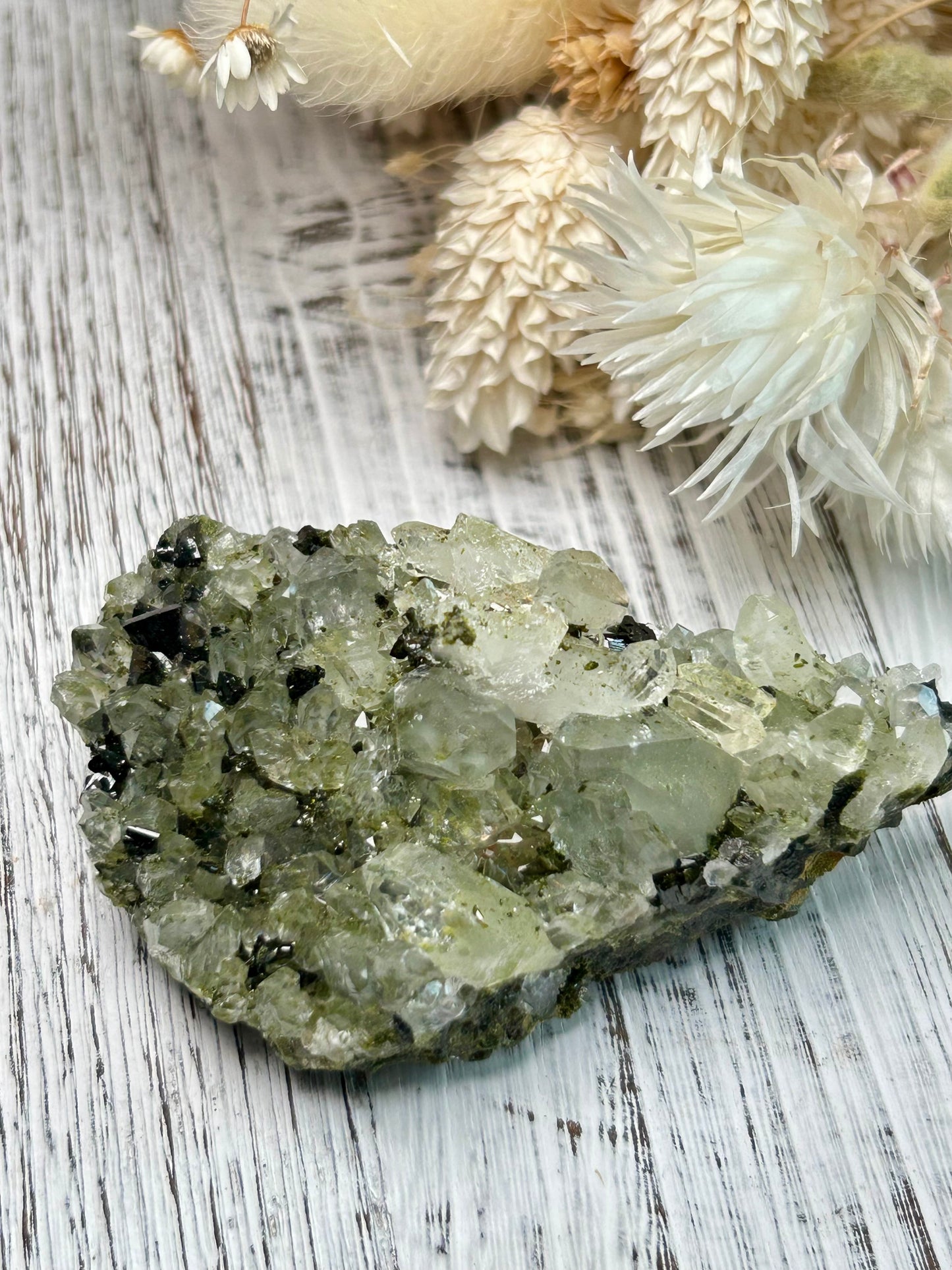 Clear quartz crystal with deep green epidote inclusions, polished freeform displayed on a distressed white wooden board with dried cream flowers