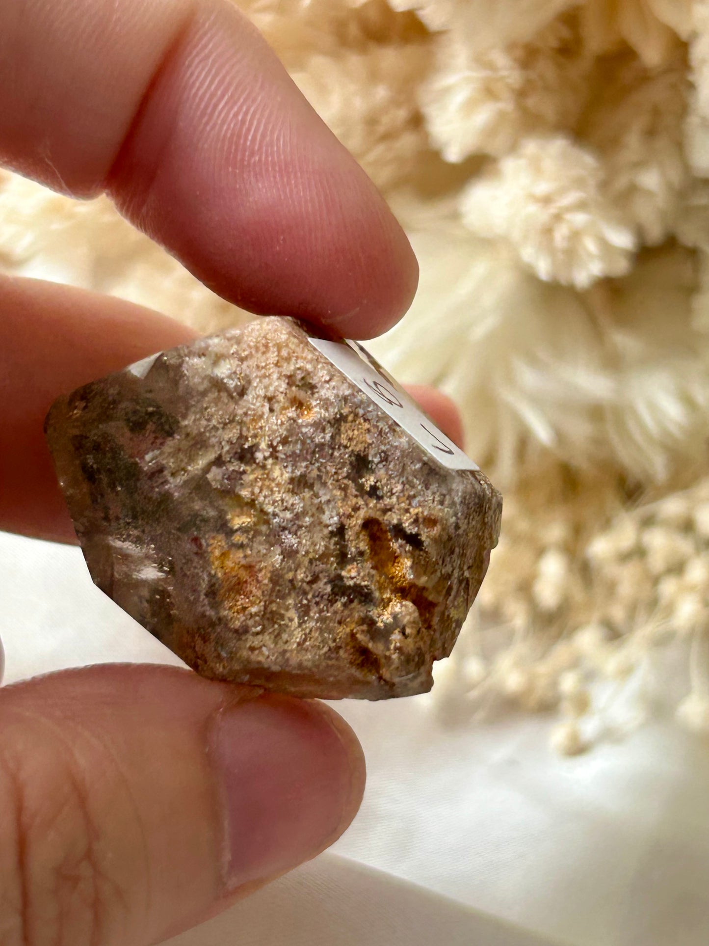 holding a Lodolite freeform crystal against an ivory satin backdrop with dried cream flowers; soft natural light highlights internal inclusions.