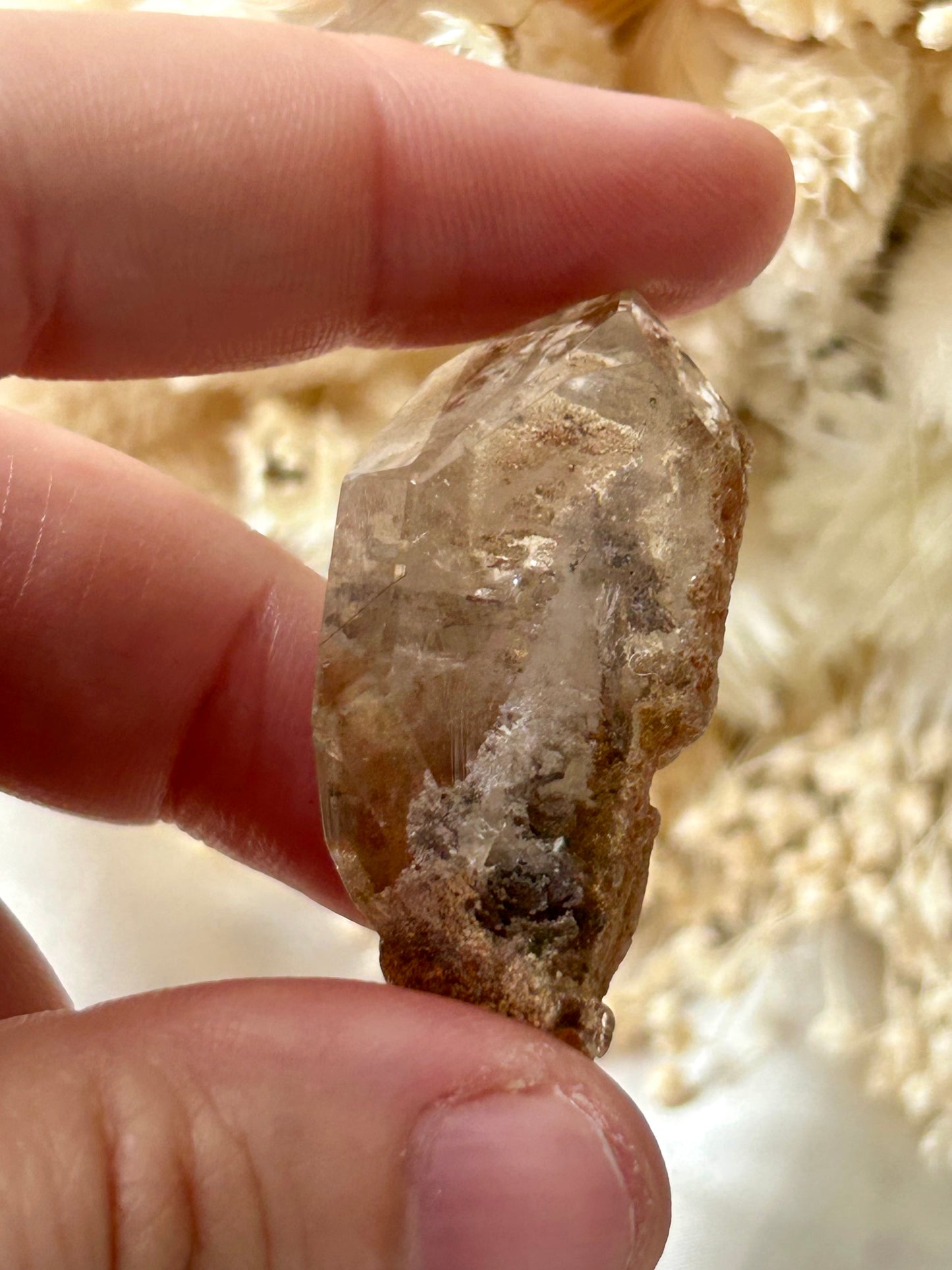 holding a Lodolite freeform crystal against an ivory satin backdrop with dried cream flowers; soft natural light highlights internal inclusions.