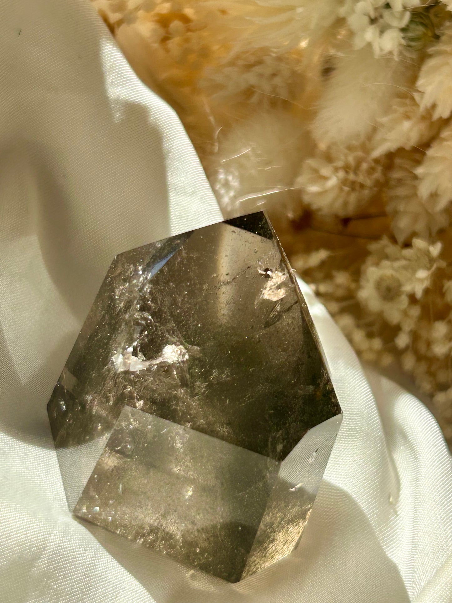 Smoky Quartz tower with chlorite inclusions resting on ivory satin, catching golden sunlight with earthy internal reflections.