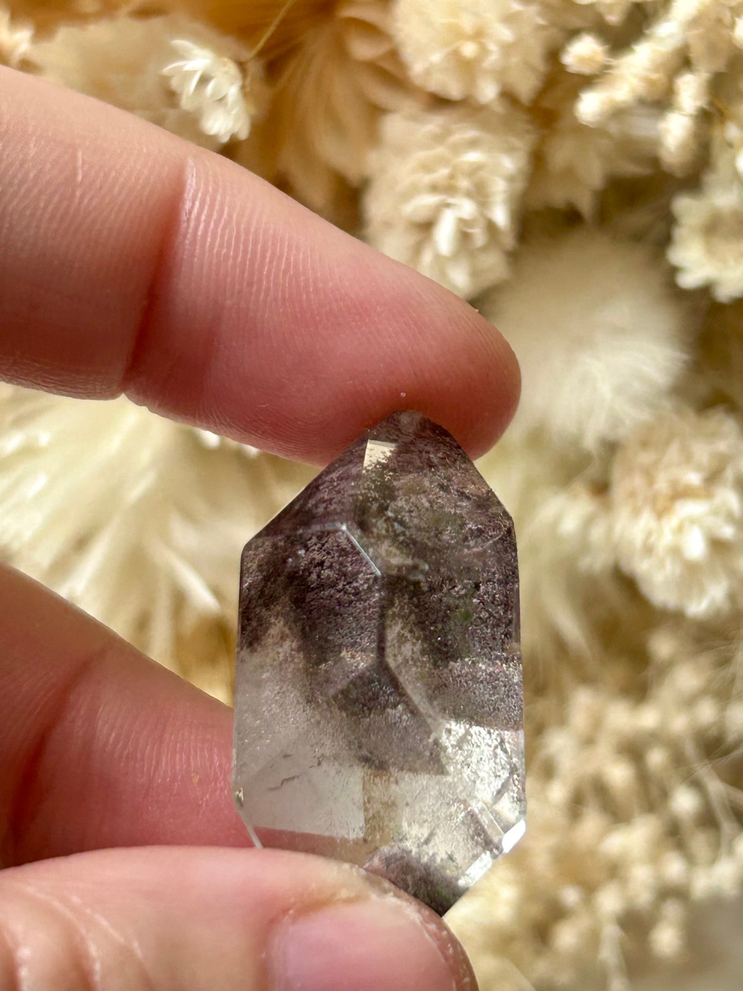 holding a Lodolite freeform crystal against an ivory satin backdrop with dried cream flowers; soft natural light highlights internal inclusions.
