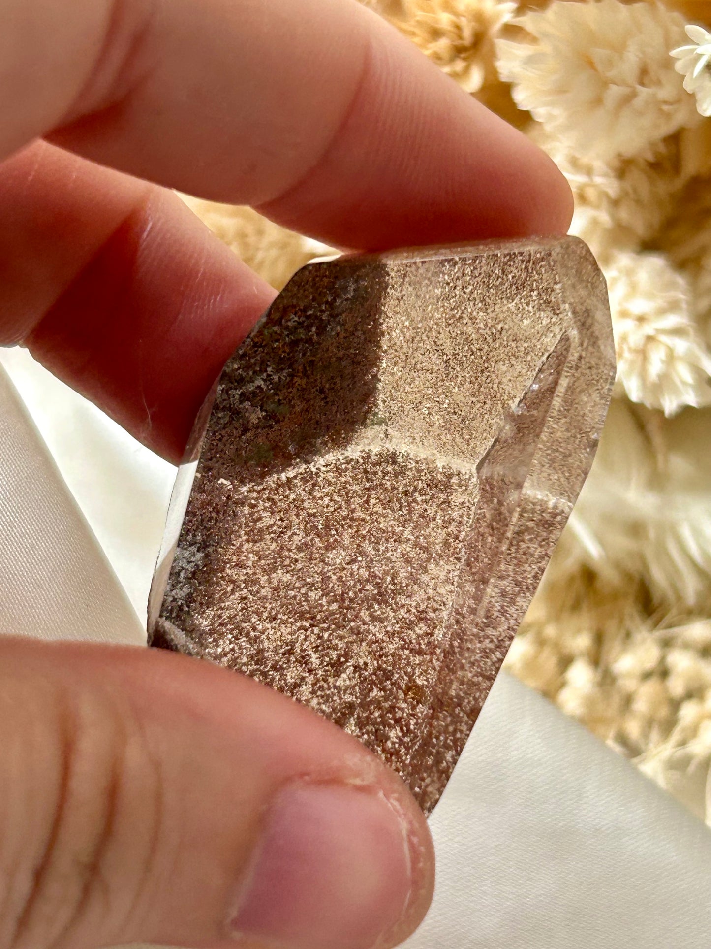 holding a Lodolite freeform crystal against an ivory satin backdrop with dried cream flowers; soft natural light highlights internal inclusions.