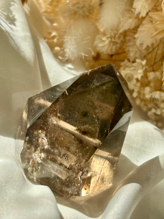 Smoky Quartz tower with chlorite inclusions resting on ivory satin, catching golden sunlight with earthy internal reflections.