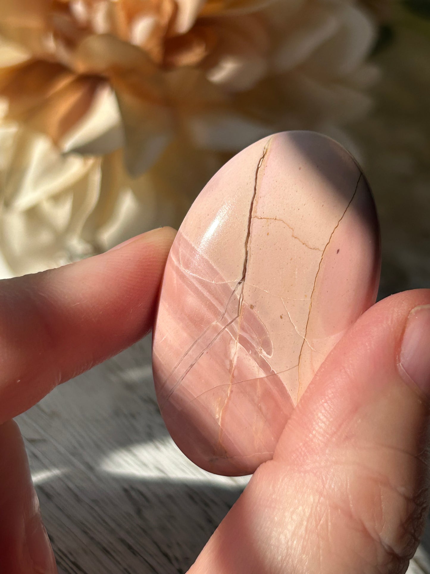 A hand holding a polished, teardrop-shaped pink stone with natural fissures, set against a soft-focus background of beige fabric and a gray textured surface.
