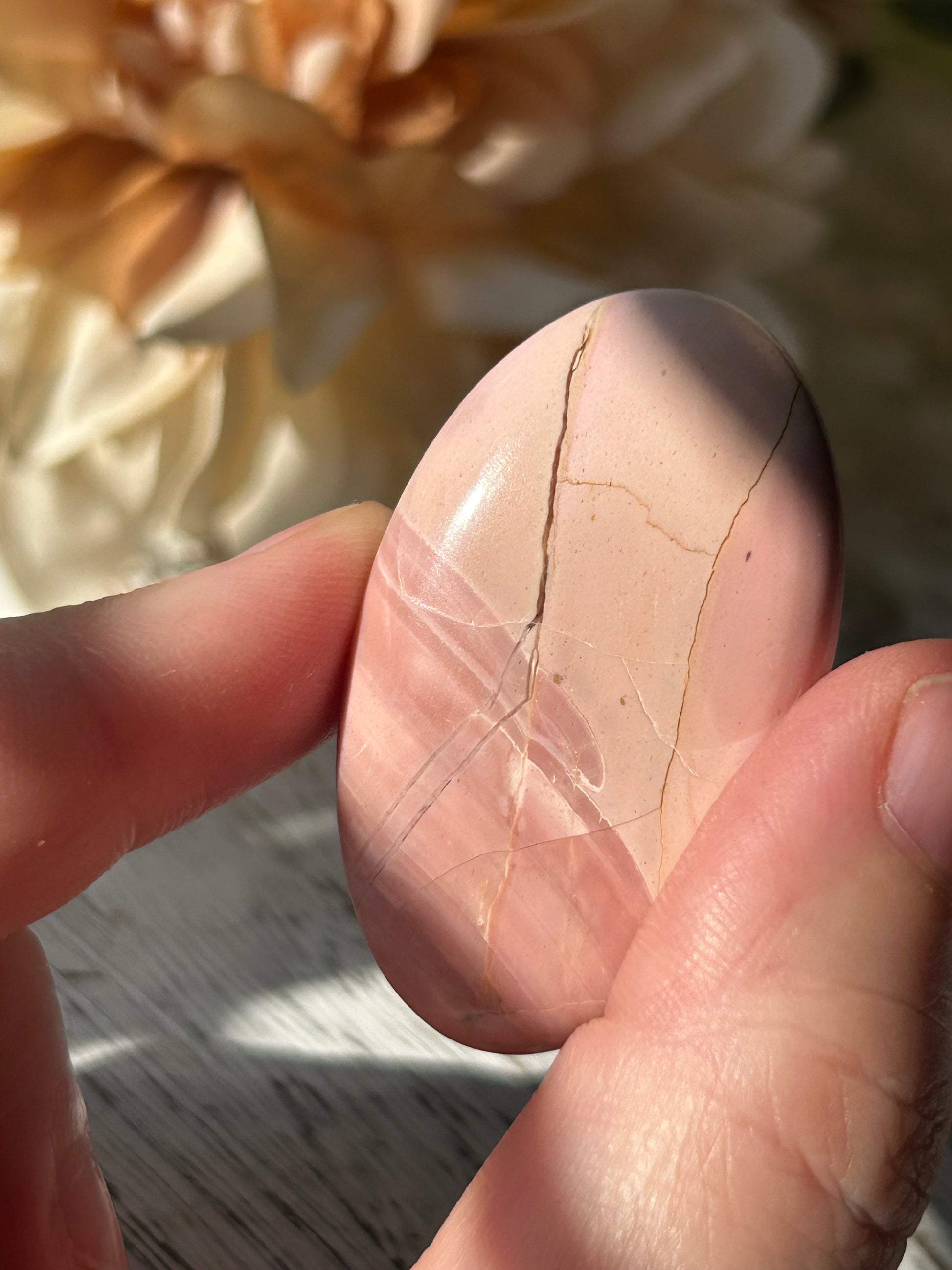 A hand holding a polished, teardrop-shaped pink stone with natural fissures, set against a soft-focus background of beige fabric and a gray textured surface.