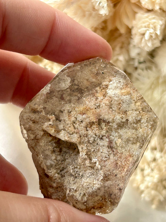 holding a Lodolite freeform crystal against an ivory satin backdrop with dried cream flowers; soft natural light highlights internal inclusions.
