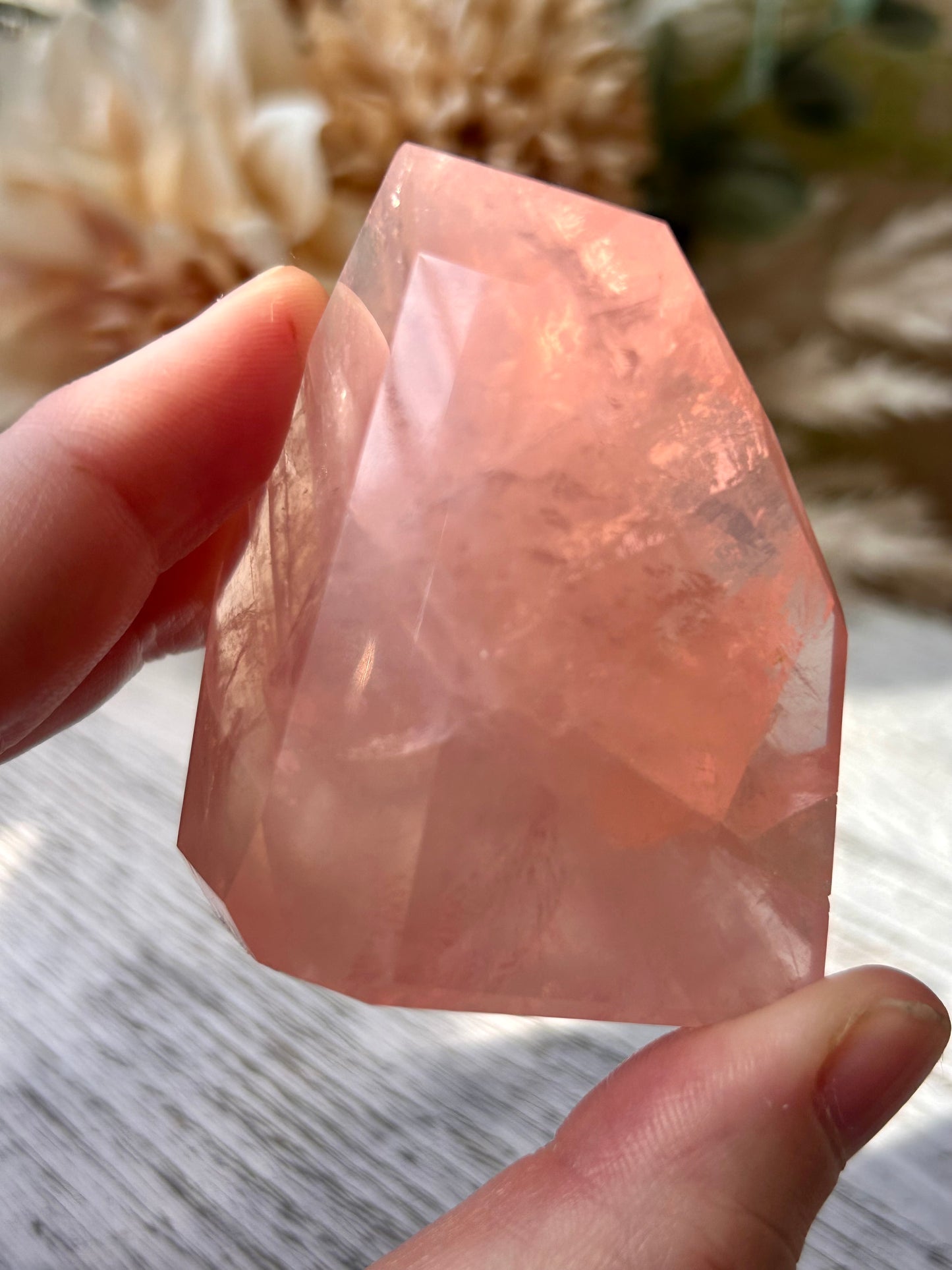 A faceted pink rose quartz crystal held between two fingers to show its shape and translucent color, with a soft-focus background of dried flowers and a light wooden surface.