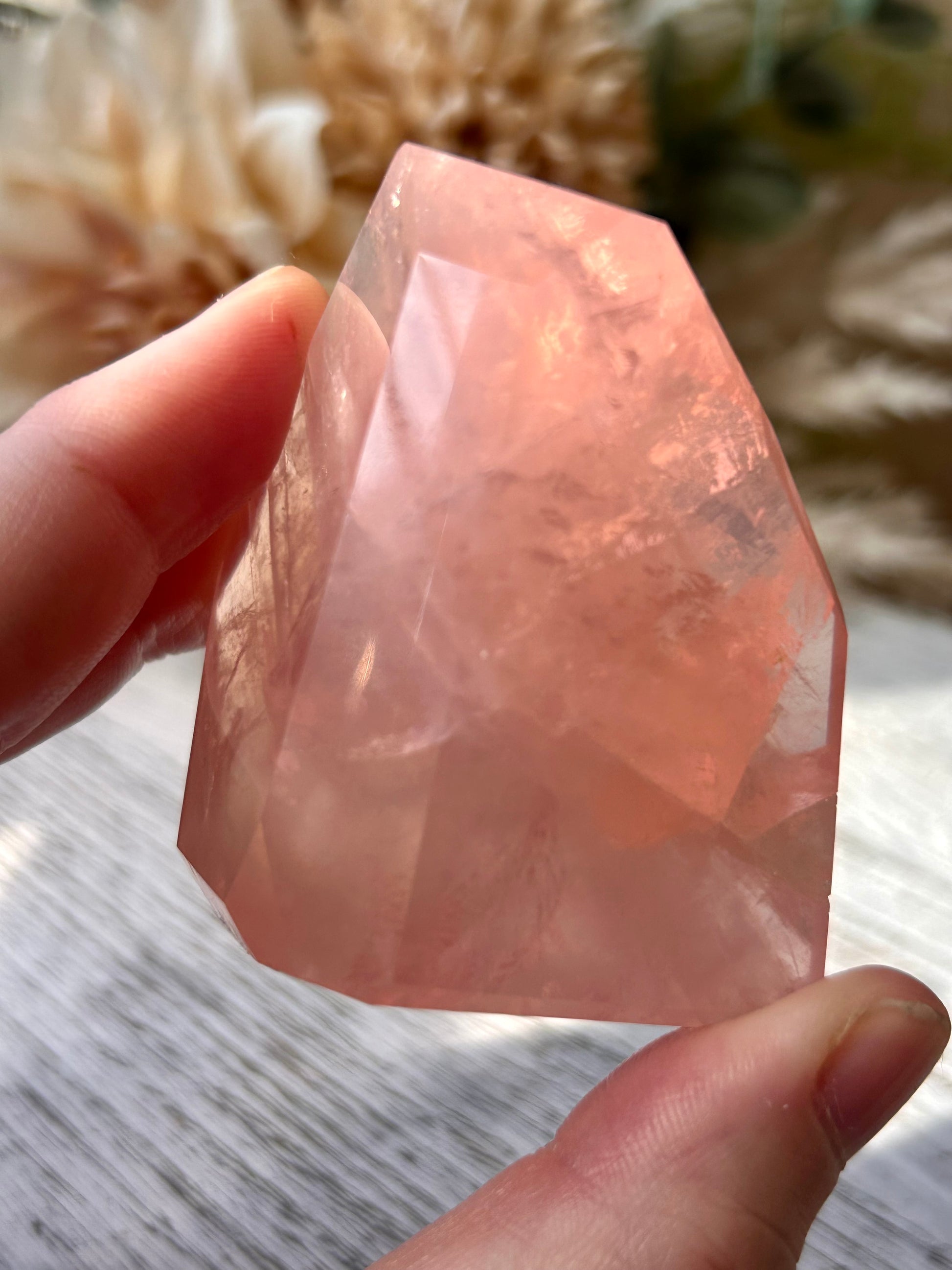 A faceted pink rose quartz crystal held between two fingers to show its shape and translucent color, with a soft-focus background of dried flowers and a light wooden surface.