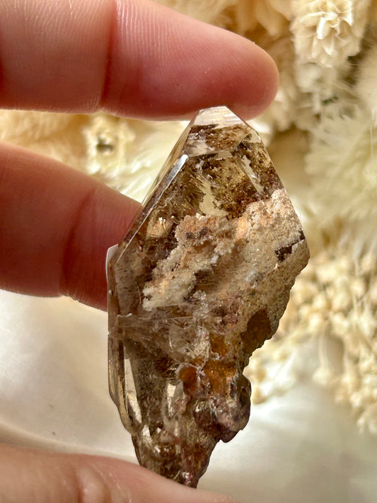 holding a Lodolite freeform crystal against an ivory satin backdrop with dried cream flowers; soft natural light highlights internal inclusions.