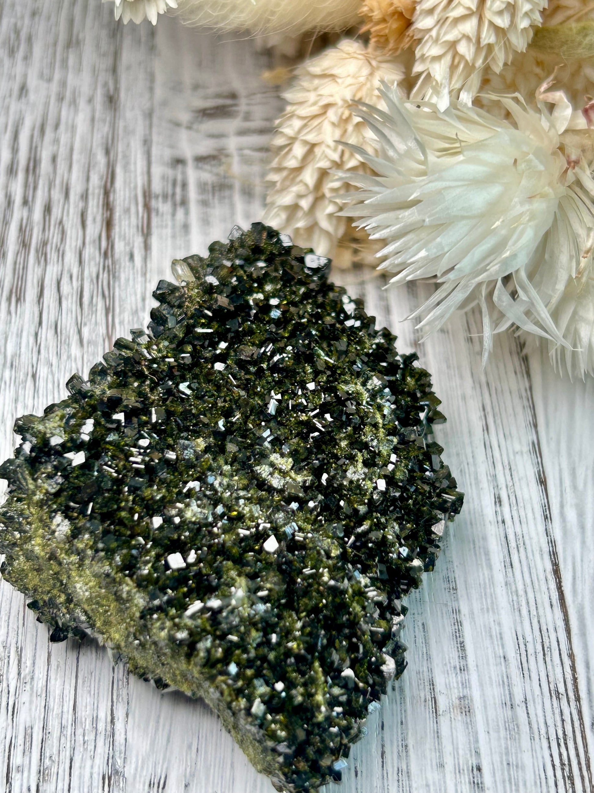 Clear quartz crystal with deep green epidote inclusions, polished freeform displayed on a distressed white wooden board with dried cream flowers 