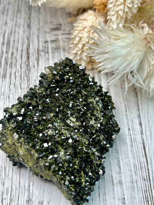 Clear quartz crystal with deep green epidote inclusions, polished freeform displayed on a distressed white wooden board with dried cream flowers 
