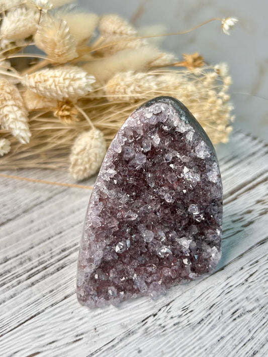 Amethyst cut base from Uruguay with vibrant violet crystal points and polished edges, displayed on a rustic white stand beside dried florals.