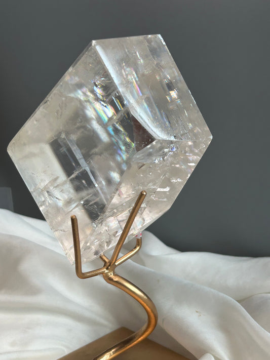 Clear Optical Calcite crystal on gold display stand showing strong light refraction and internal rainbows against a soft white fabric background.