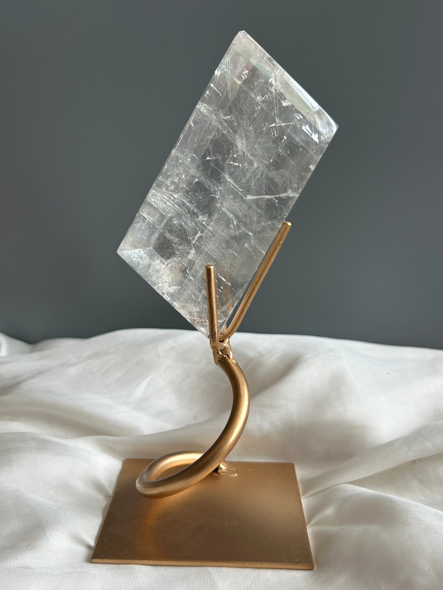 Clear Optical Calcite crystal on gold display stand showing strong light refraction and internal rainbows against a soft white fabric background.