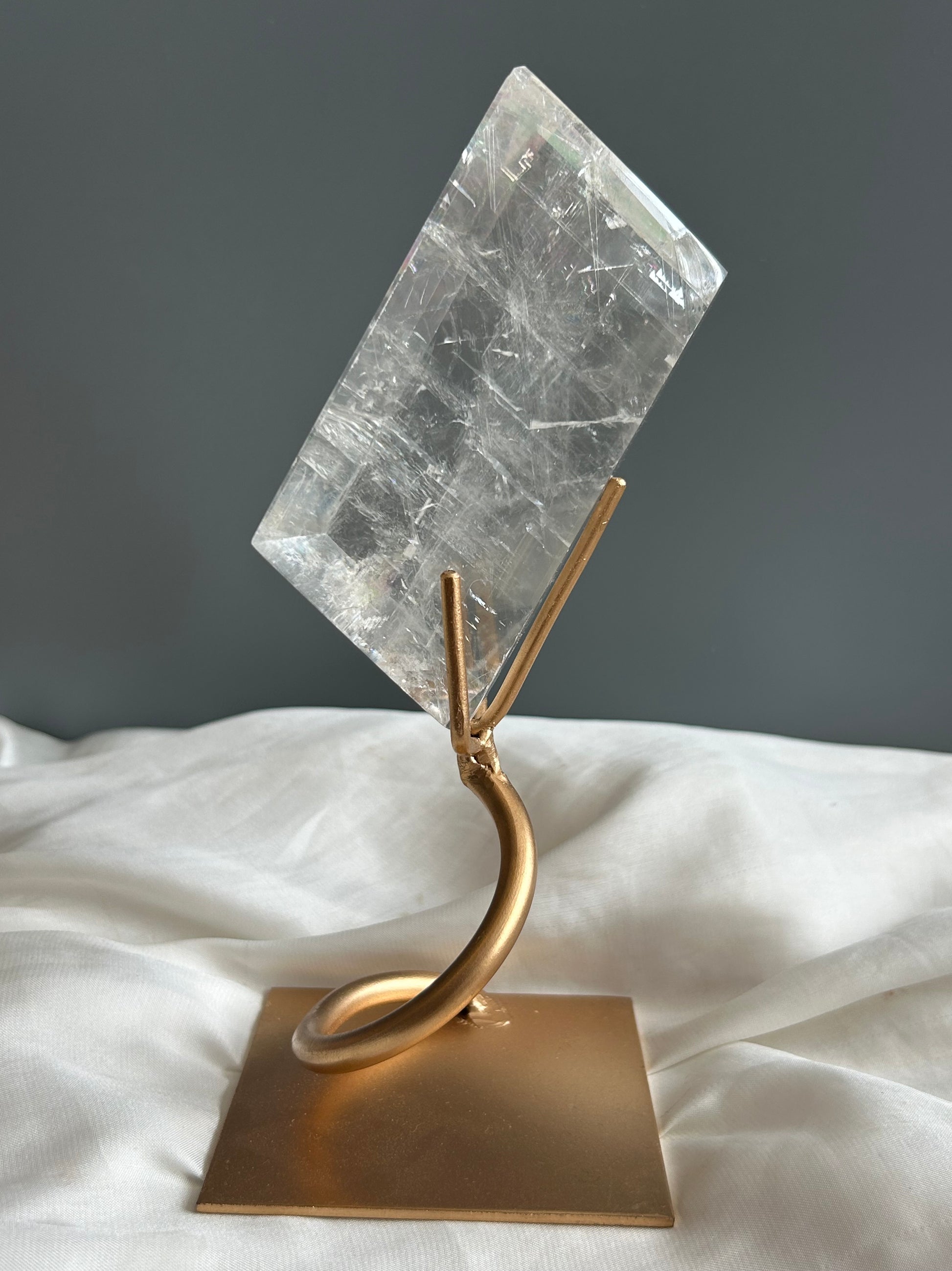 Clear Optical Calcite crystal on gold display stand showing strong light refraction and internal rainbows against a soft white fabric background.