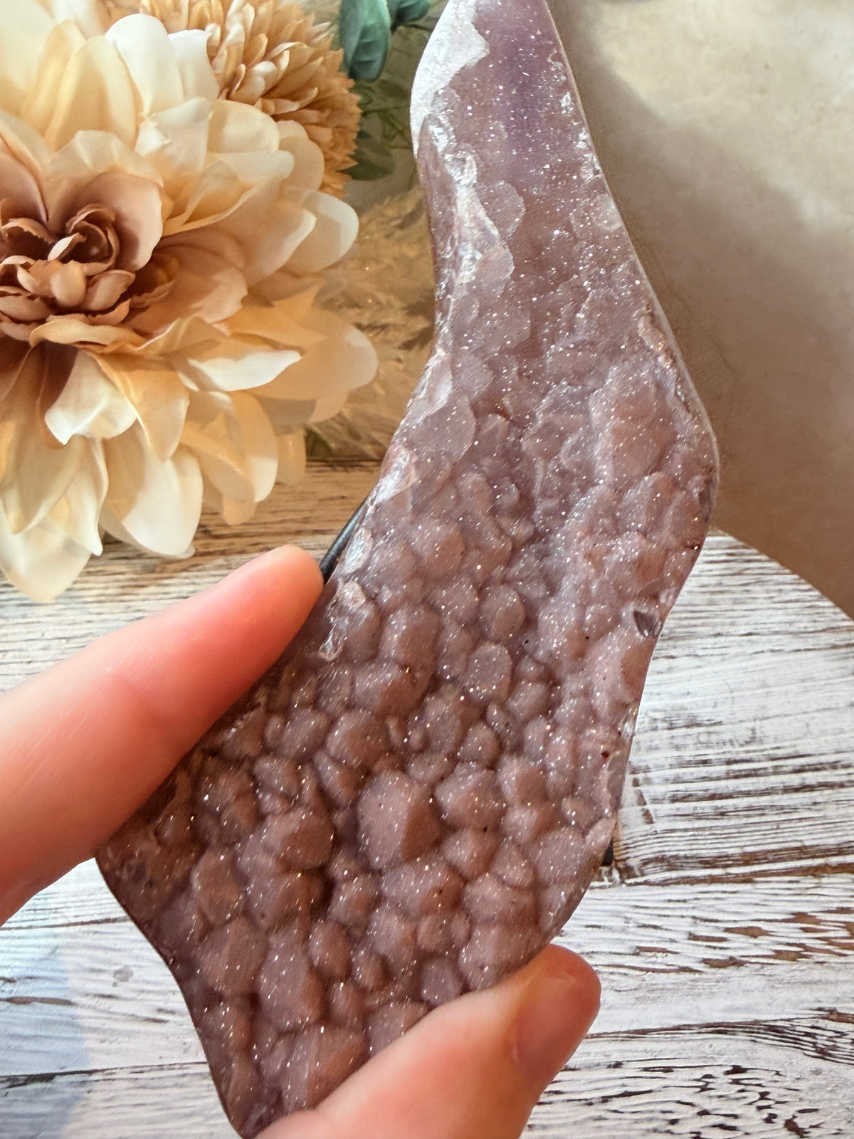 A hand holding a polished irregular-shaped agate or quartz crystal with a textured, bubble-like internal structure. The crystal has a dusty lavender-purple color with fine glitter.