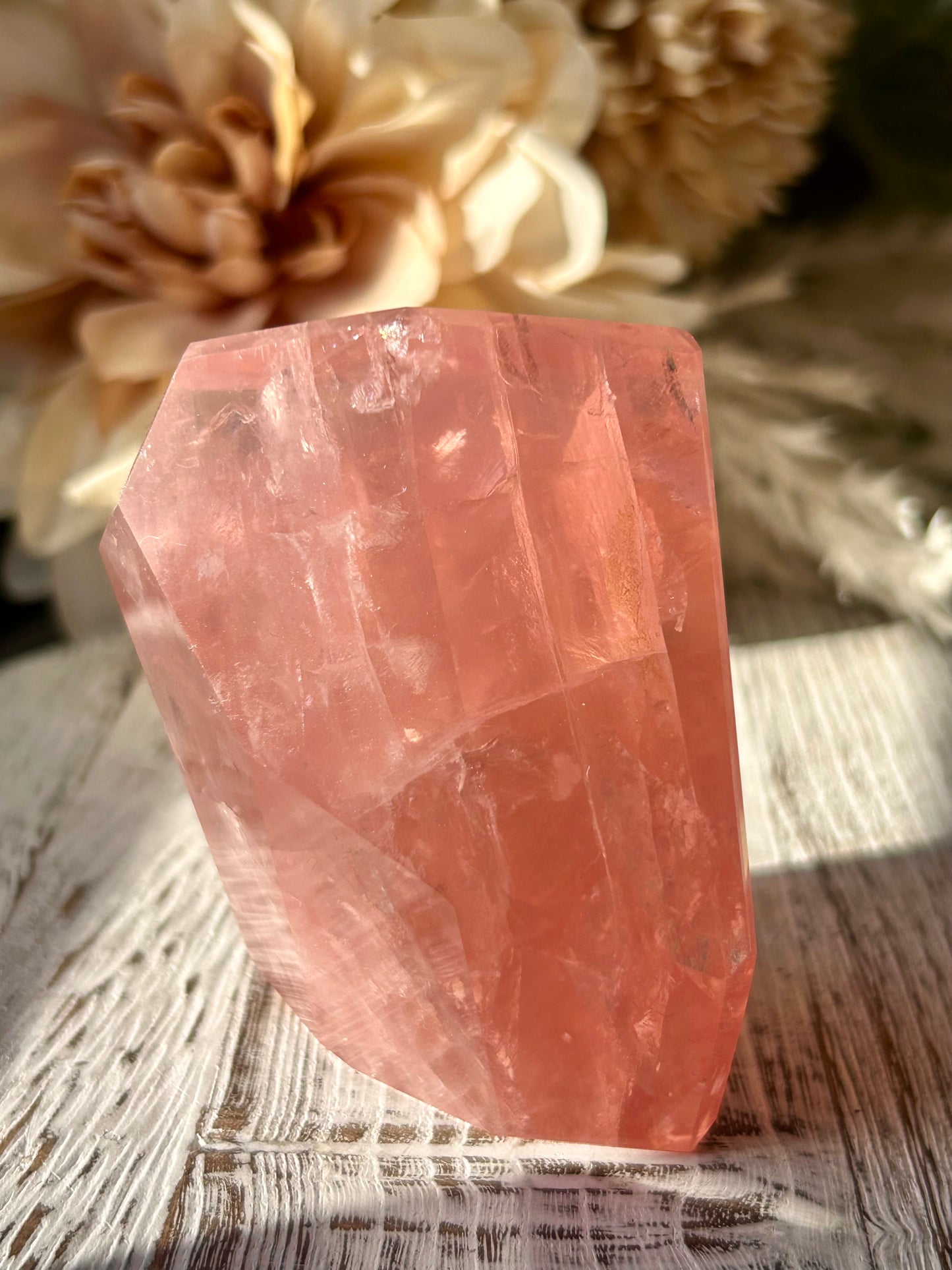 A faceted pink crystal stone, likely rose quartz, standing on a rustic white-washed wooden surface with soft beige dried flowers blurred in the background.