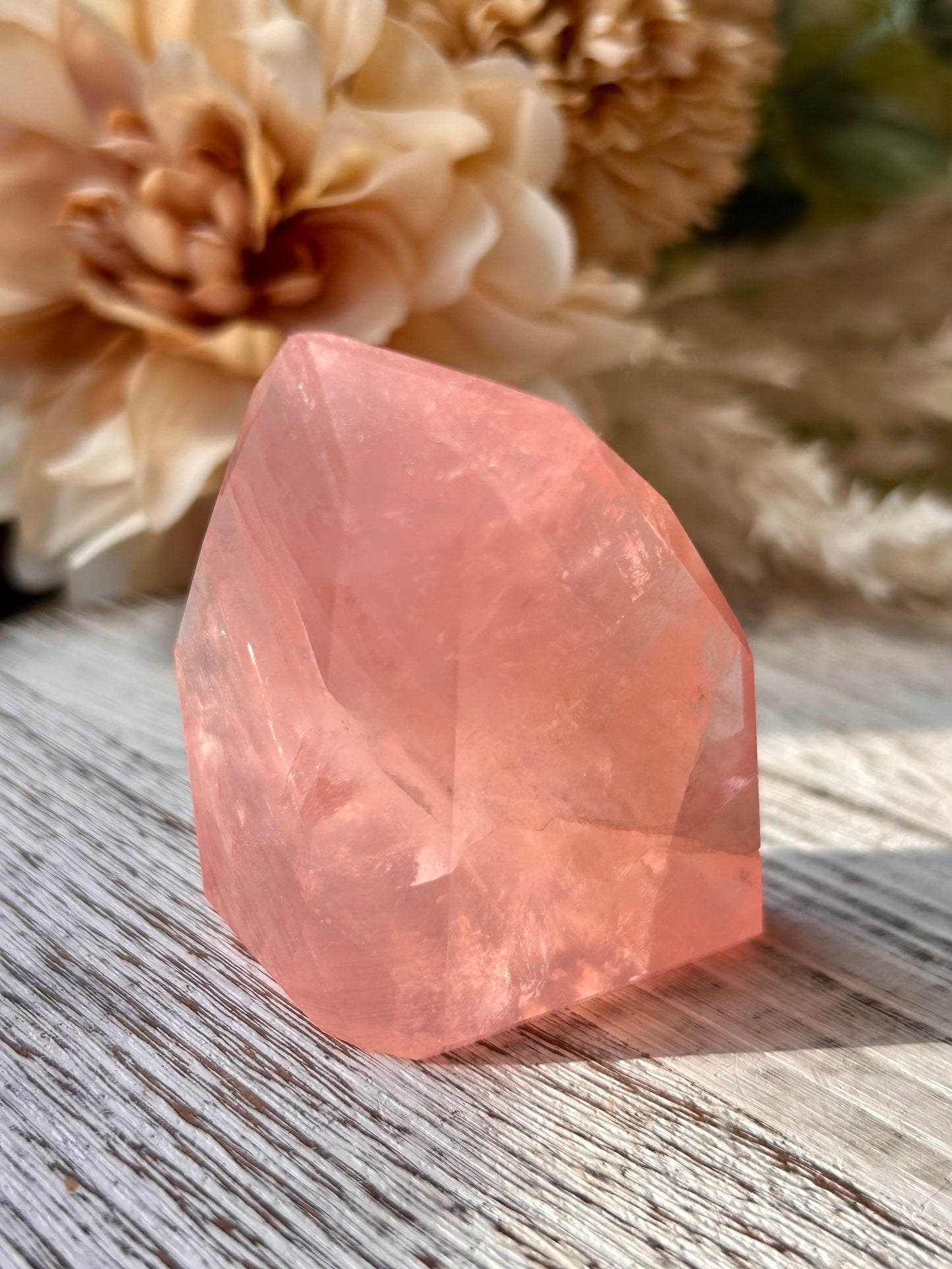 A faceted pink crystal stone, likely rose quartz, standing on a weathered gray wooden surface with blurred dried flowers in the background.