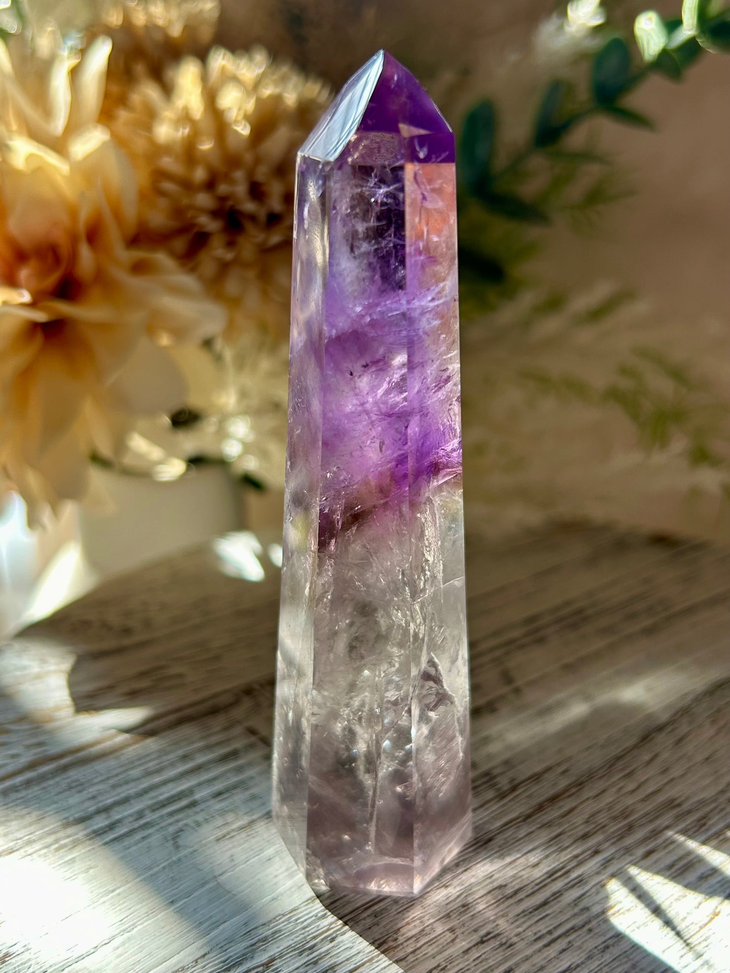 Crystal tower with purple and clear sections on a wooden surface with flowers in the background