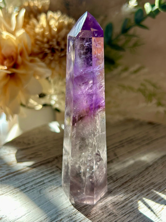 Crystal tower with purple and clear sections on a wooden surface with flowers in the background