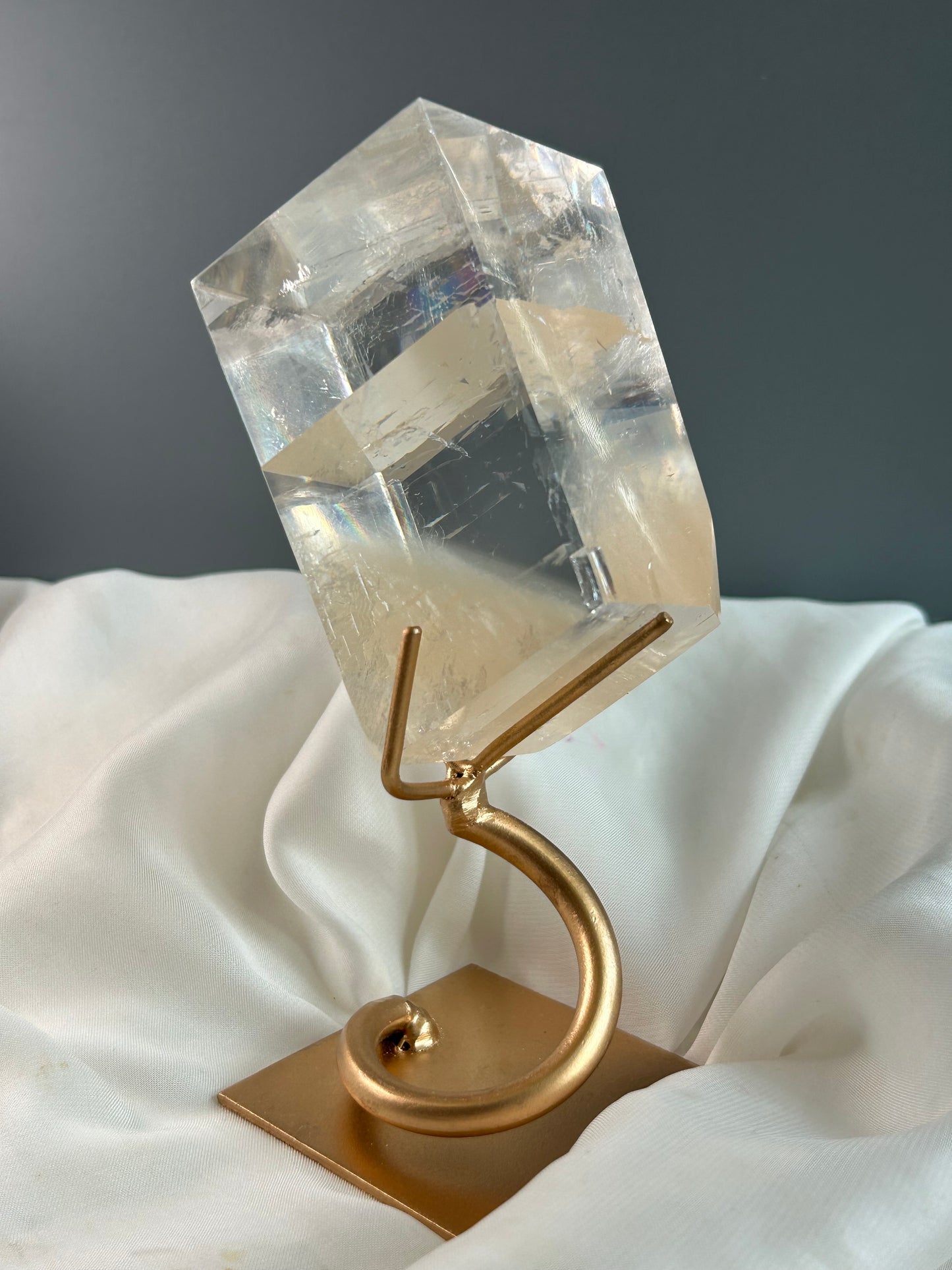 Clear Optical Calcite crystal on gold display stand showing strong light refraction and internal rainbows against a soft white fabric background.