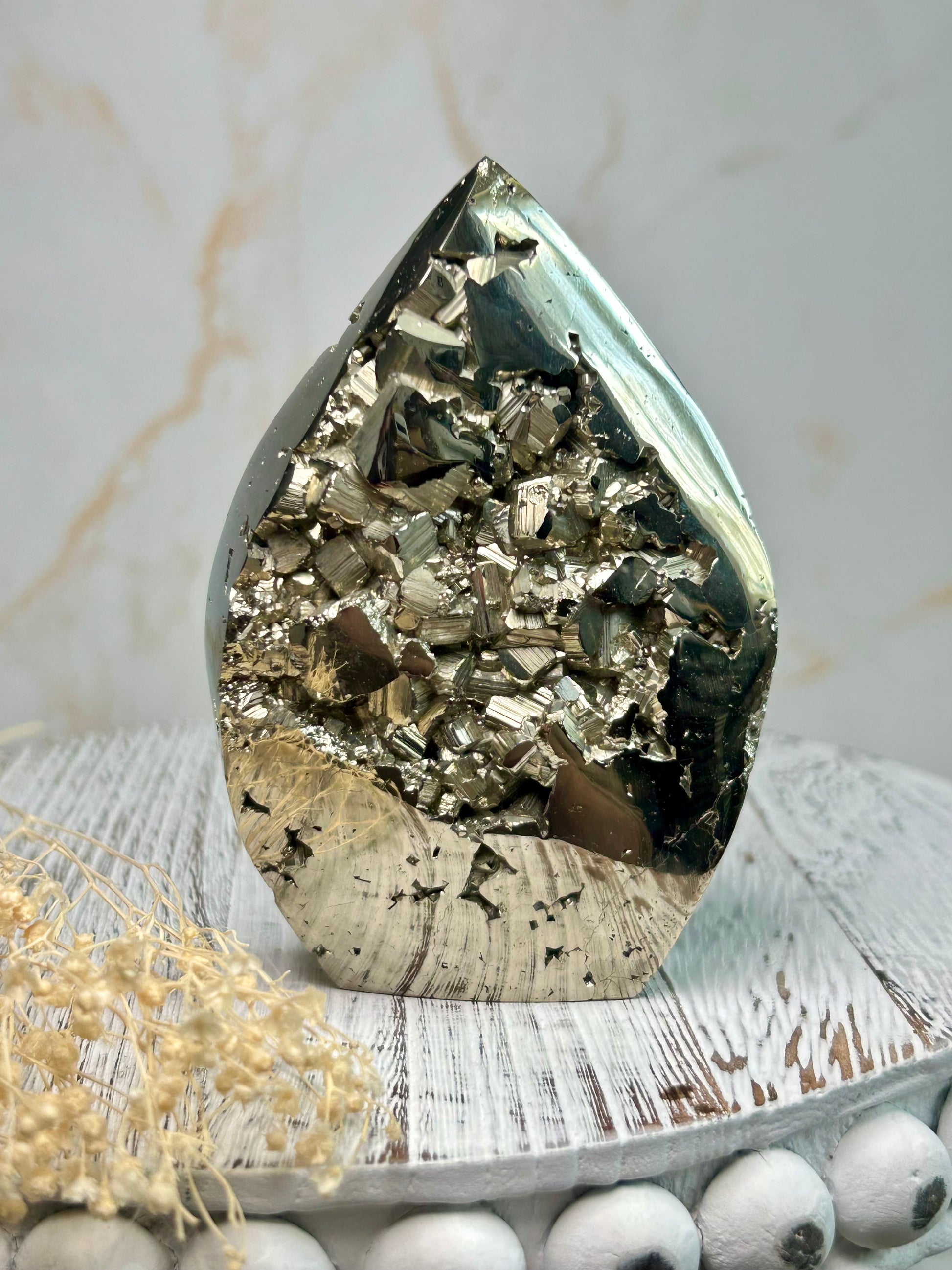 Golden pyrite flame crystal polished into flame shape, shimmering metallic surface displayed on a distressed wooden board with cream dried flowers.