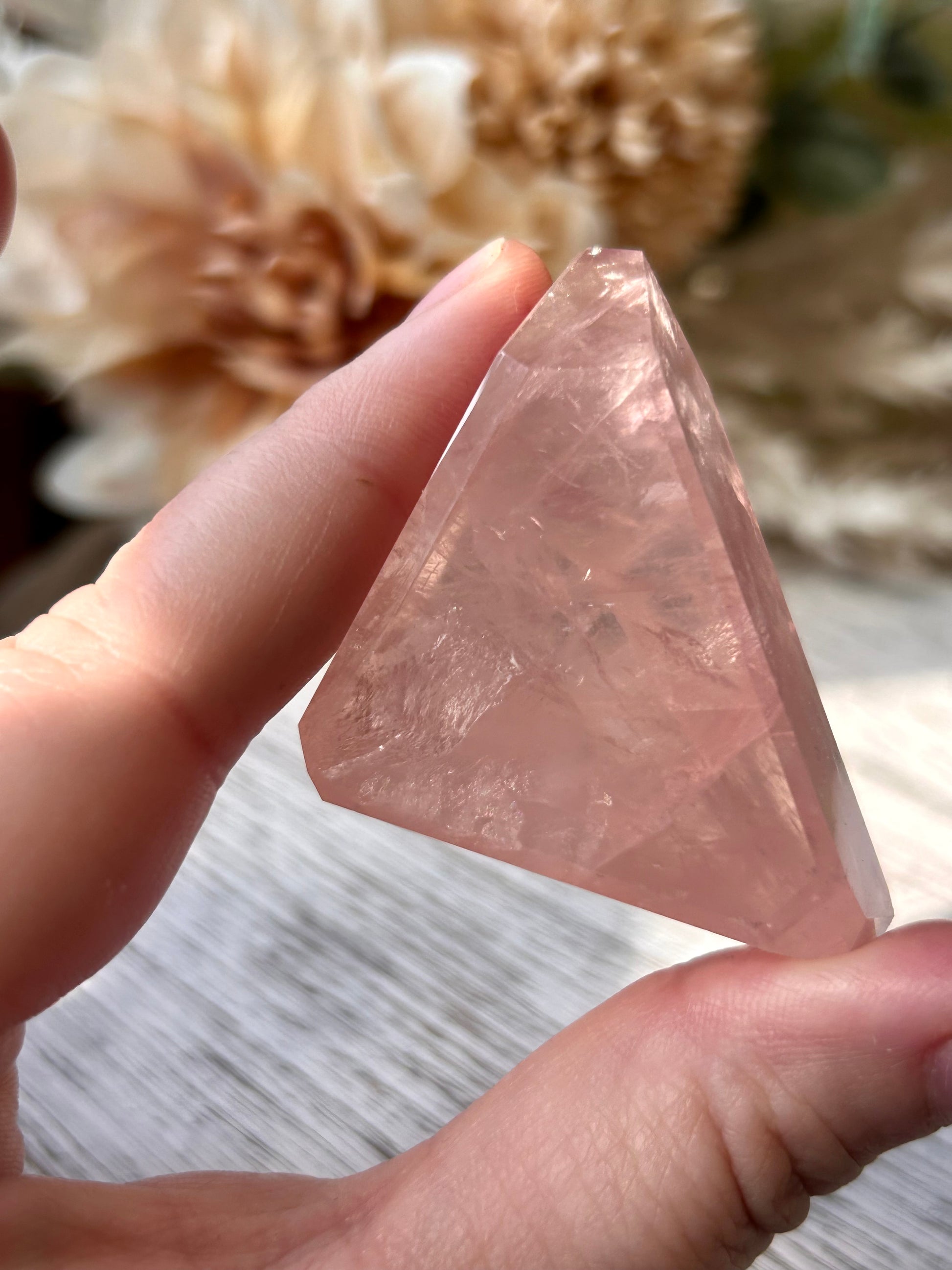 A hand holding a faceted pink crystal pyramid with natural inclusions, set against a soft-focus background of dried flowers and a light wood surface.
