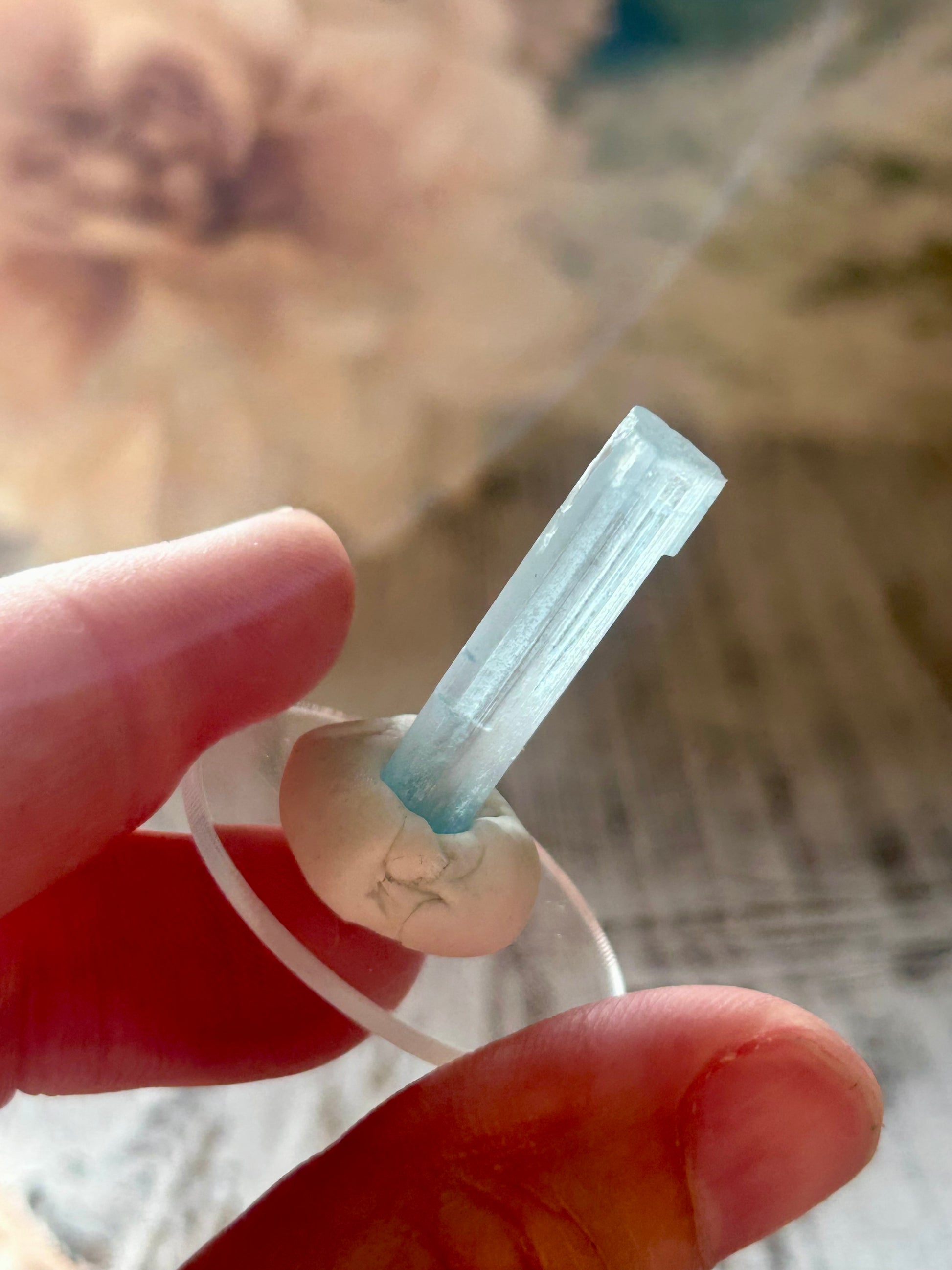 A hand holding a small clear cylindrical crystal or glass vial with a pale blue liquid inside, against a softly blurred beige background.