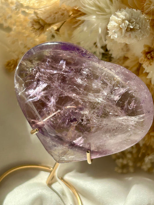 Polished Amethyst heart crystal from Brazil with a deep purple phantom inclusion and visible rainbow flashes, resting on ivory satin with dried flowers in the background