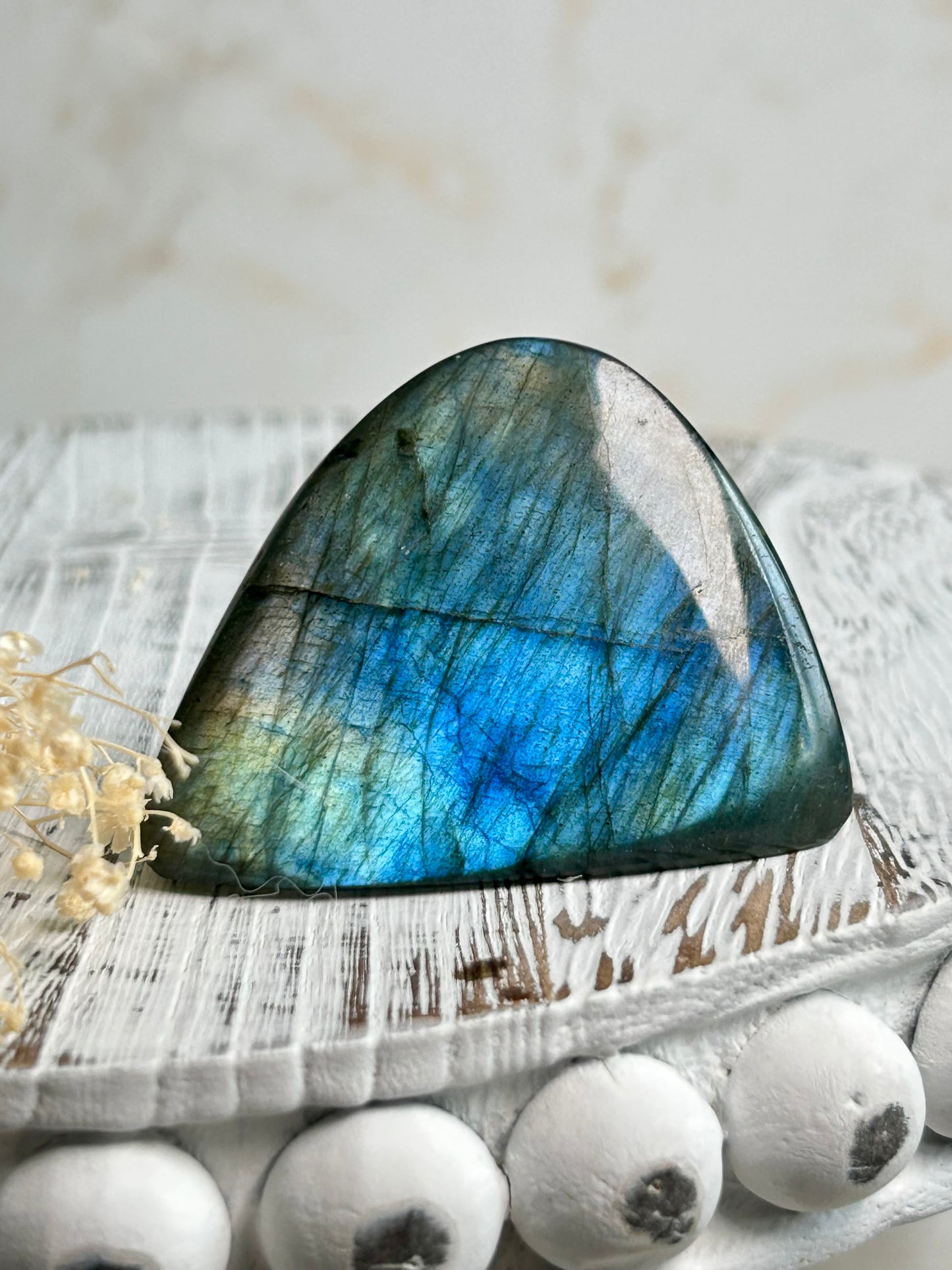 Polished labradorite freeform with vivid blue flash and iridescent surface on a distressed white wooden board with dried cream flowers
