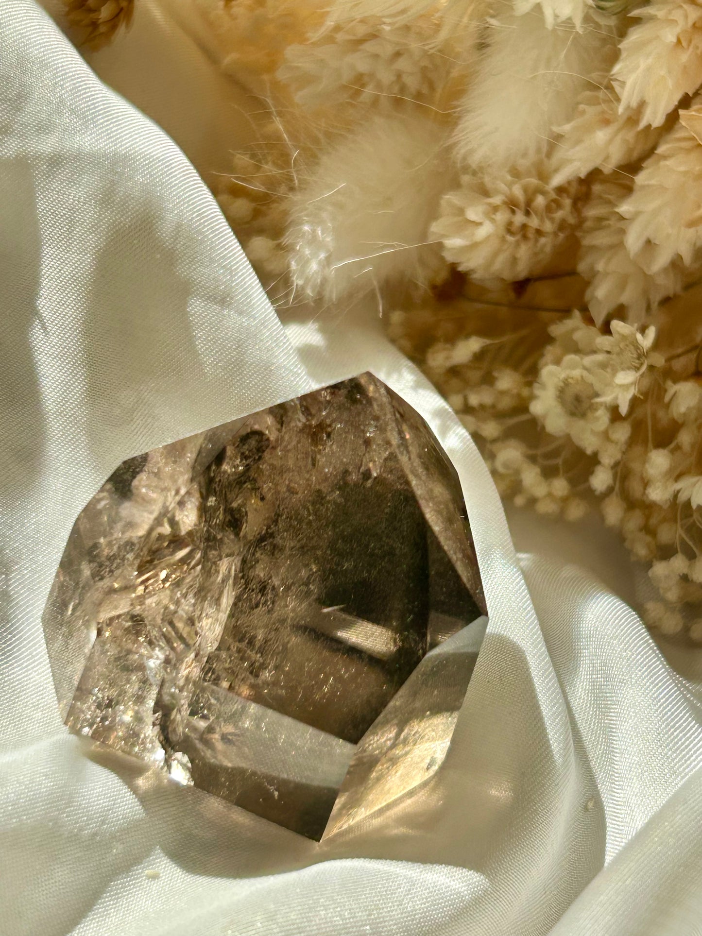 Smoky Quartz tower with chlorite inclusions resting on ivory satin, catching golden sunlight with earthy internal reflections.