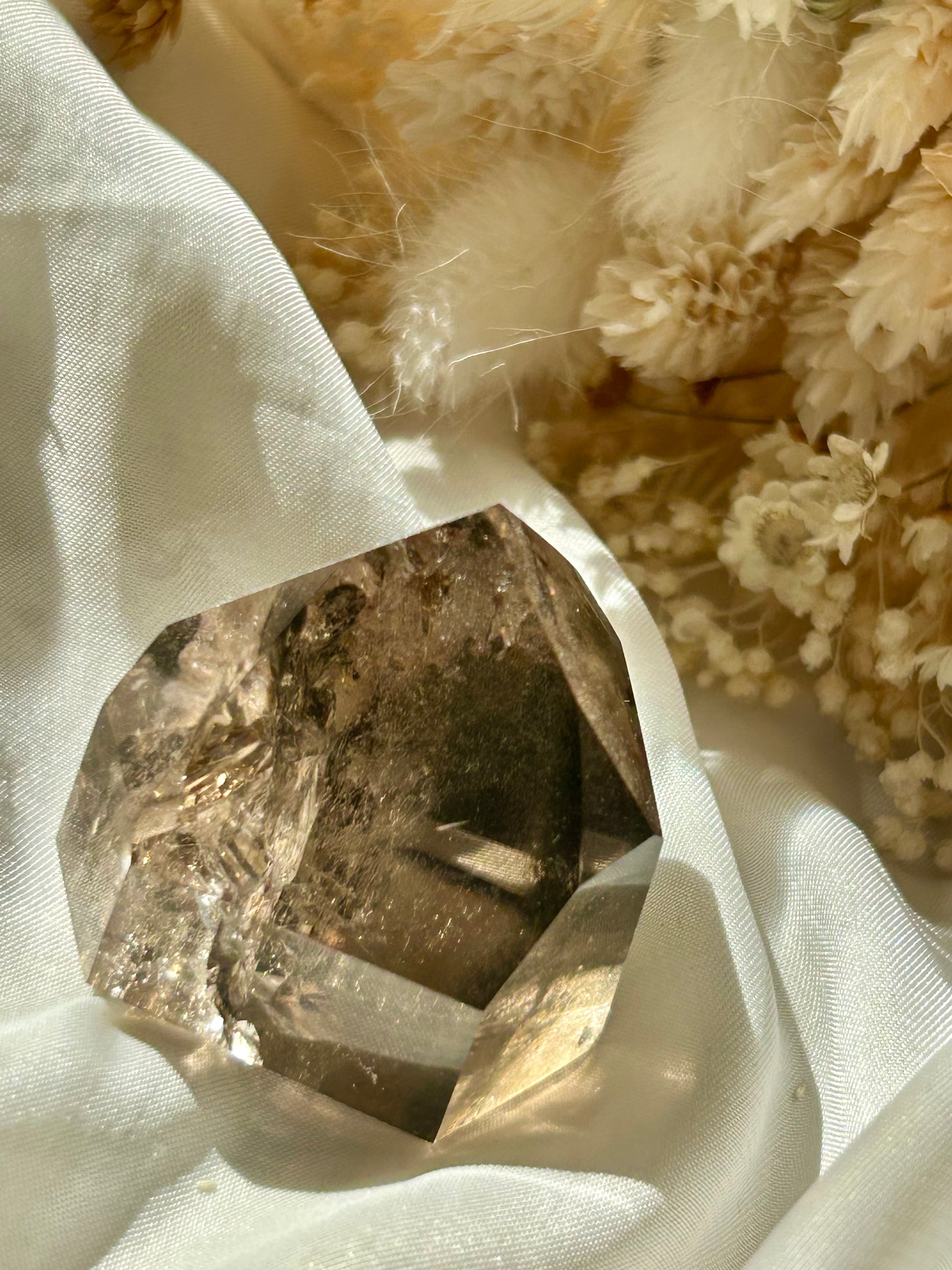 Smoky Quartz tower with chlorite inclusions resting on ivory satin, catching golden sunlight with earthy internal reflections.