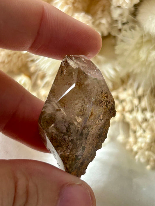holding a Lodolite freeform crystal against an ivory satin backdrop with dried cream flowers; soft natural light highlights internal inclusions.