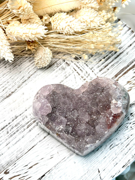 Lilac Sugar Amethyst heart from Uruguay with soft sparkling druzy surface, displayed on a rustic white surface beside dried florals.