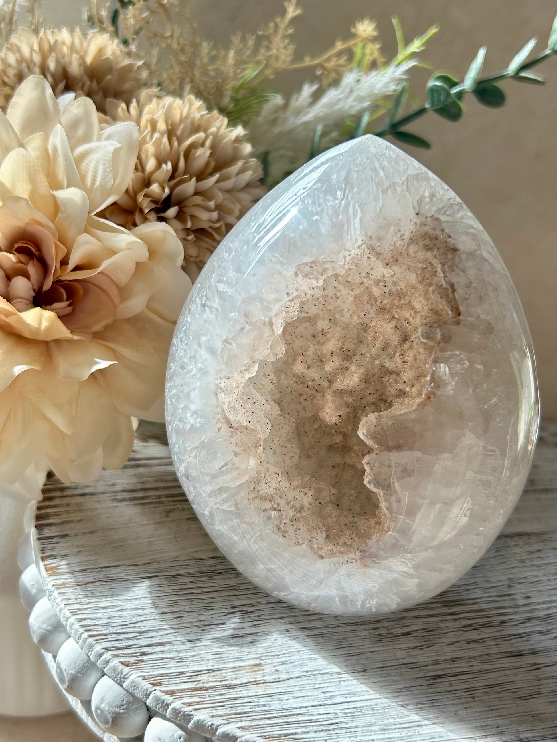 Polished pale druzy crystal freeform with a sparkling natural cavity at the centre, photographed in warm sunlight beside soft cream dried flowers on a rustic whitewashed surface.