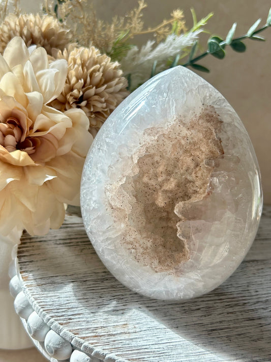 Polished pale druzy crystal freeform with a sparkling natural cavity at the centre, photographed in warm sunlight beside soft cream dried flowers on a rustic whitewashed surface.