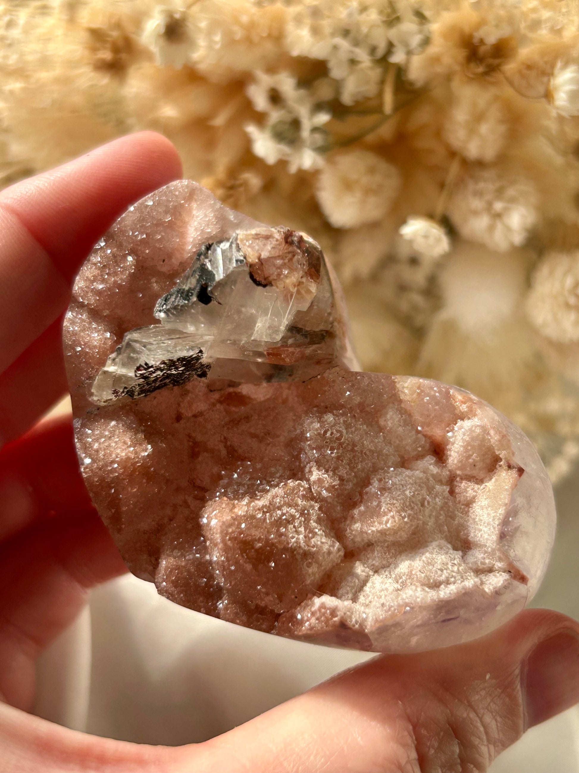 Blush pink druzy Amethyst heart with exposed mineral inclusions and raw crystal clarity, held in hand with dried floral background
